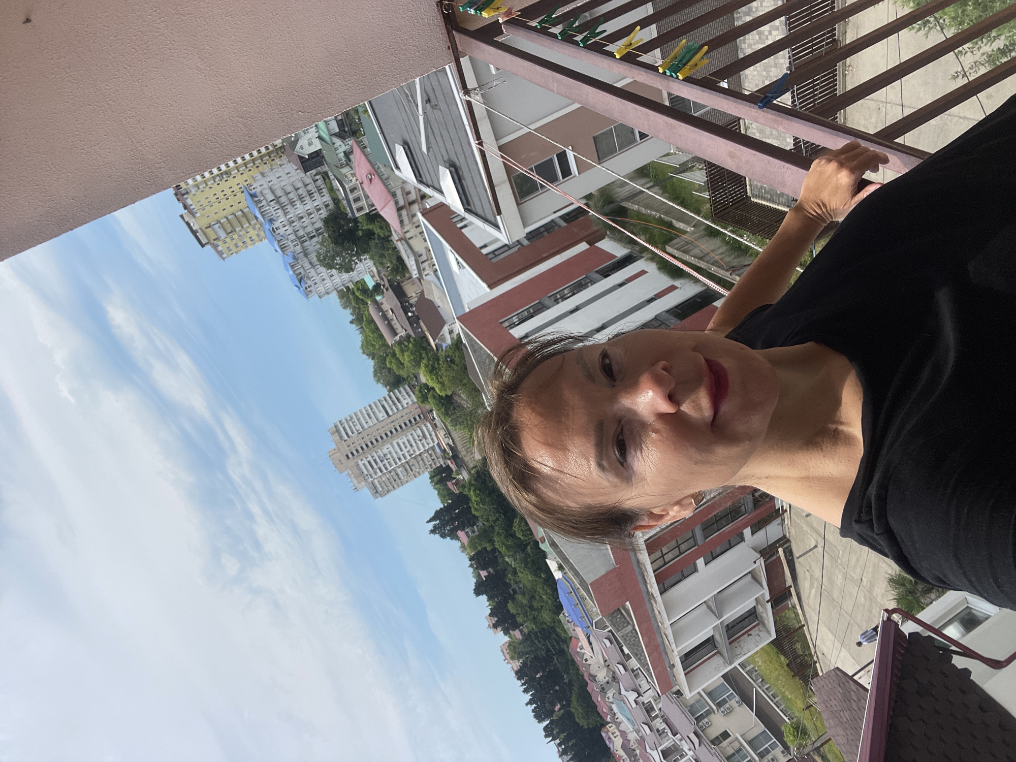 An overhead, angled, selfie-style photograph taken from a balcony on a sunny day in Sochi, Russia. The image features a woman in the foreground wearing a black t-shirt. Her right arm is resting on the balcony railing. The background shows a dense urban landscape with numerous multi-story buildings of varying architectural styles and colors, some with red roofs. Trees are interspersed between the buildings, suggesting a hilly terrain. The sky is blue with scattered white clouds. A clothesline with several clothespins is visible on the balcony railing. A person in a blue and white striped shirt is walking on a path below. The camera angle emphasizes the depth of the city stretching out into the distance.