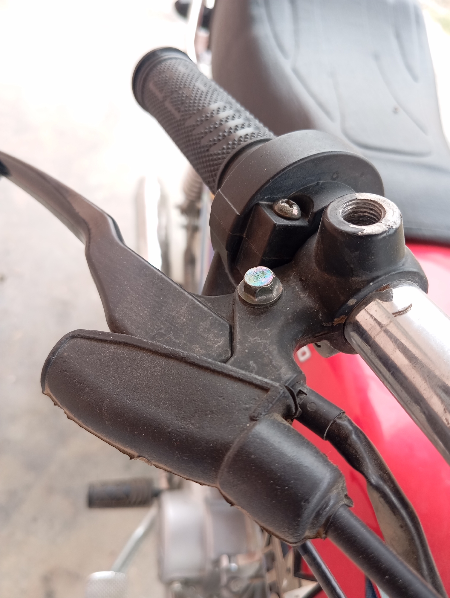 A close-up shot shows the handlebar and front brake lever of a red motorcycle. The brake lever is black and appears to be made of plastic. It is mounted on a black housing attached to the silver handlebar. A bolt with a holographic sheen secures the brake lever assembly. To the right of the brake lever, a threaded metal fixture is visible, possibly for attaching a cable or accessory. The textured grip of the handlebar is also visible, as is a portion of the motorcycle's red body. The background is out of focus, suggesting an outdoor setting.