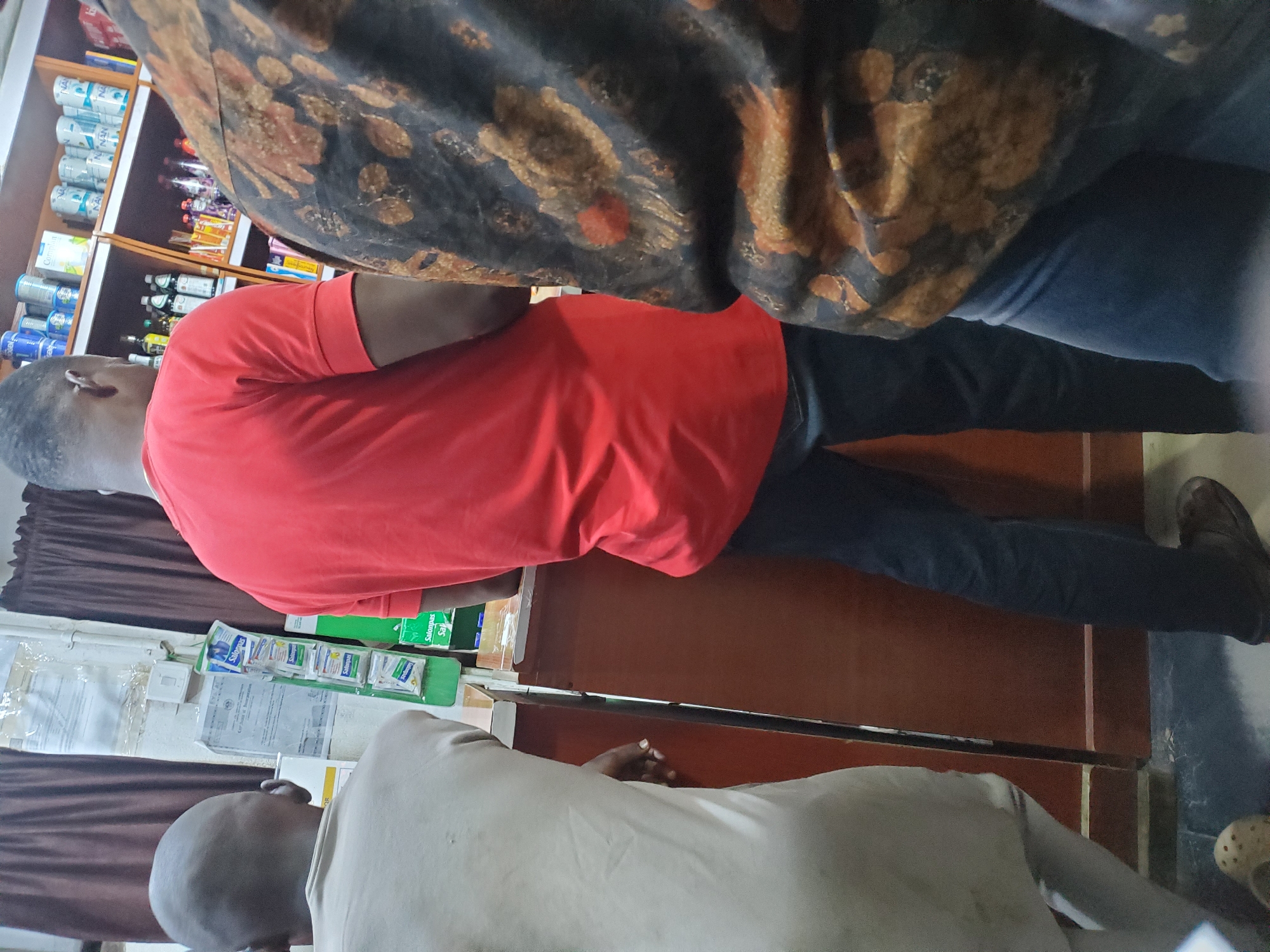 The image depicts a scene inside a shop in Jos, Nigeria. Three individuals are partially visible. On the left, a person wearing a light-colored garment stands facing away from the camera. In the center, a man in a red t-shirt and dark trousers stands facing away. To the right, another person's torso is visible, wearing a dark patterned shirt over blue trousers. Shelves stocked with various items, including canned goods and bottles, are visible in the background. A display rack with small packaged items, some labeled "Salonpas," is also present. The environment appears to be a retail setting, possibly a convenience store or pharmacy.