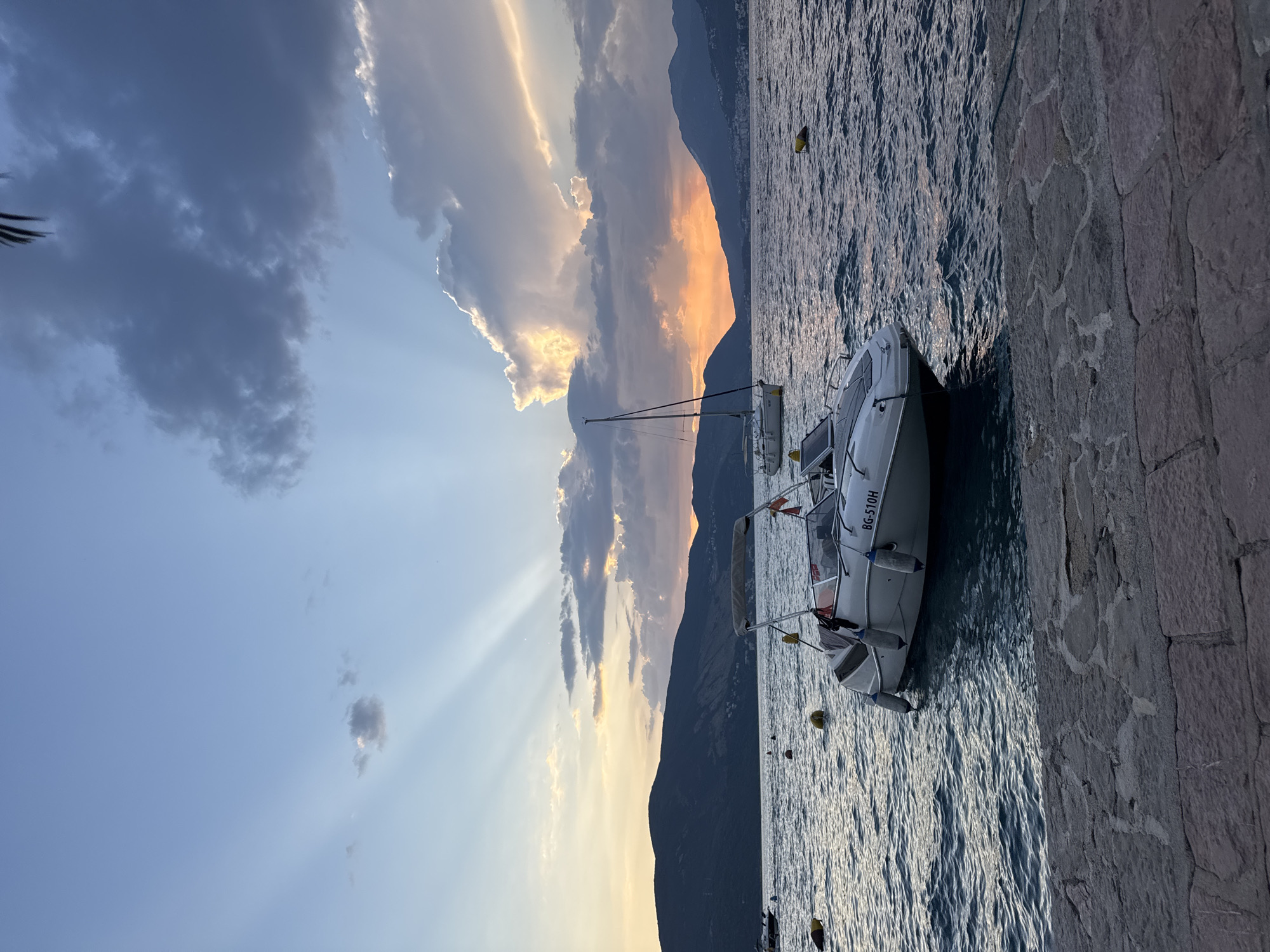 A boat is moored to a stone embankment by a body of water. The boat is white with a dark red trim and has a registration number that reads "BG-510H". There are several yellow buoys floating in the water. In the background, dark mountains rise from the water's edge and extend into the distance. The sky is filled with dramatic clouds, some of which are illuminated by the setting sun, casting a warm orange glow. Sunbeams are visible, radiating from behind the clouds. The overall scene evokes a sense of tranquility and the beauty of a sunset.