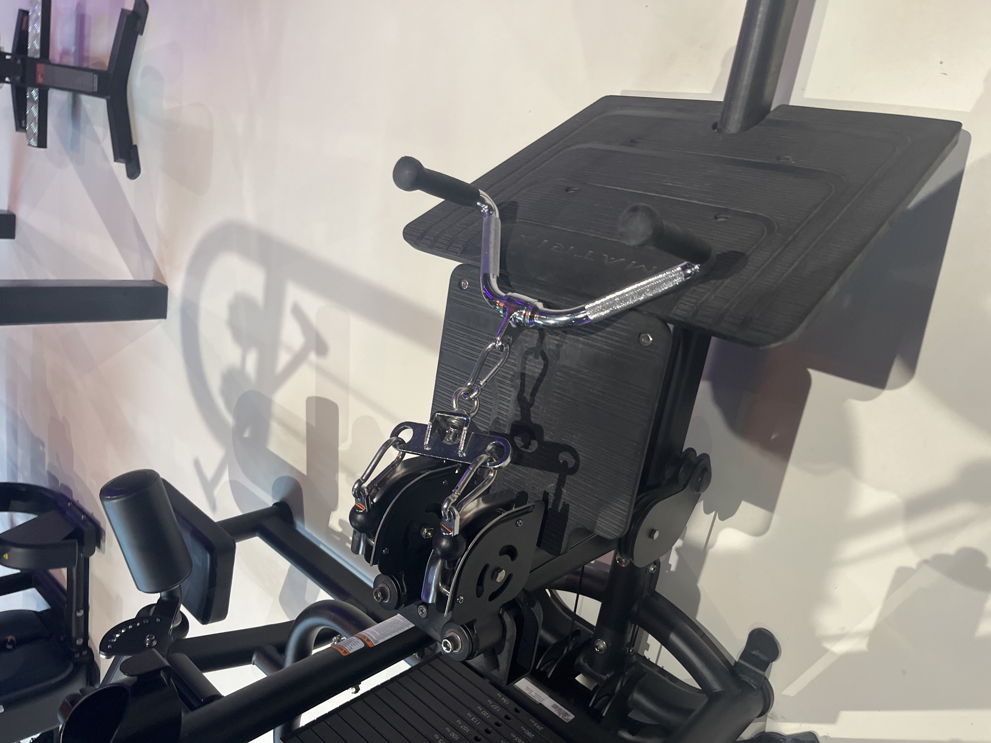 The image depicts a black exercise machine, possibly located indoors against a light-colored wall. The machine features a platform with a handle, pulleys, and what appears to be a seat with a padded support. The machine's components are constructed from metal and some type of plastic material. Other exercise equipment is visible in the background, with a rack-like structure mounted on the wall. Shadows are visible on the wall. The location and city details of Moscow, Russia are not evident in the image.