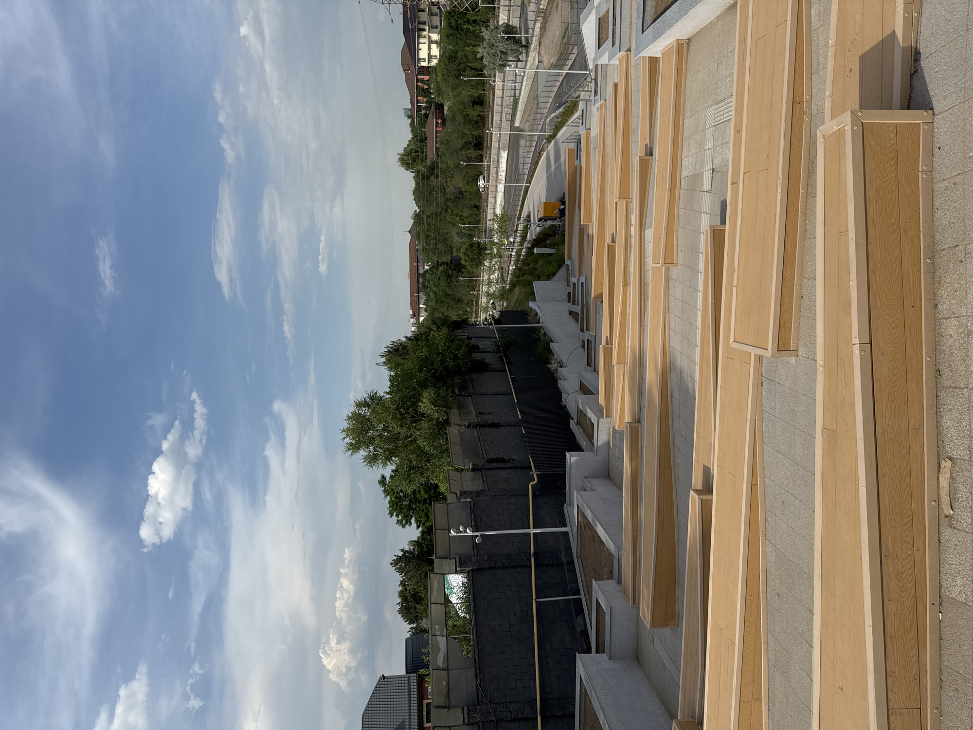 The image depicts an outdoor amphitheater with tiered wooden seating arranged on a paved surface. The seating structures are primarily made of light brown wood with contrasting lighter wooden edges. In the background, a dark, brick-like wall with fencing runs horizontally across the frame, separating the amphitheater from a road and some residential buildings. Trees and lush greenery are visible beyond the wall, and a white car is seen on the road. The sky is blue with scattered white clouds. The scene appears to be in a modern urban park or public space, likely in Almaty, Kazakhstan, given the context. There are no people visible in the image. The lighting suggests it is daytime.