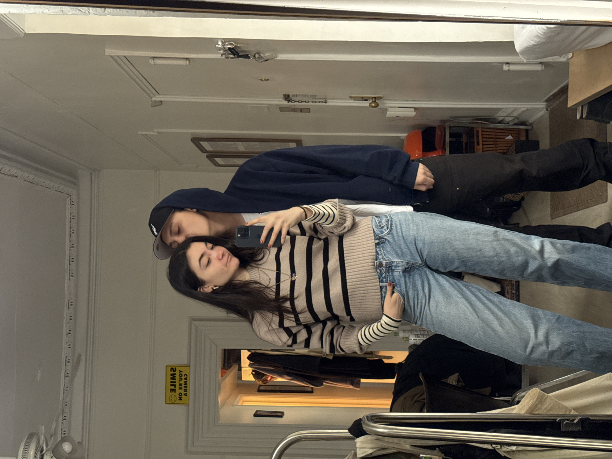 Two people are reflected in a mirror. One person is wearing a navy blue hooded sweatshirt and a baseball cap. The other person is wearing a beige and black striped sweater and blue jeans. The scene appears to be inside a room with white walls and trim, and a glimpse of a hallway can be seen in the mirror's reflection. A yellow sign with black lettering is visible on the wall. Clothes are visible on a rack in the foreground. It is not possible to confirm that this image was taken in New York, United States of America.