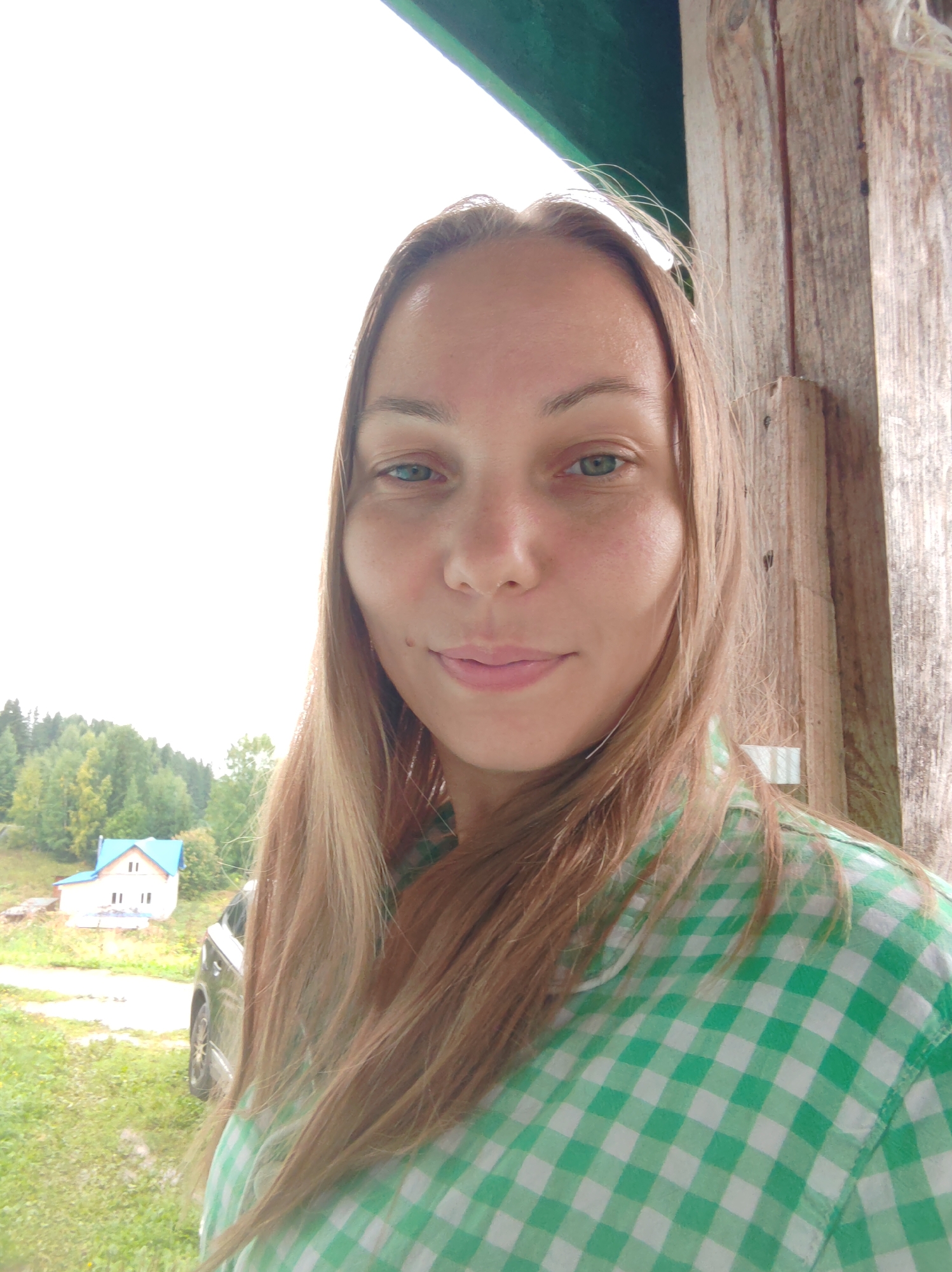A woman with long, light brown hair is taking a selfie outdoors. She is wearing a green and white checkered shirt. Her face is turned towards the camera, and she is smiling slightly. Her eyes are green. Behind her, there is a wooden structure to her right, and to her left, a bright white sky, a green forest, and a small house with a blue roof. A car is parked on a dirt road in front of the house. The scene appears to be in a rural area, possibly in or near the city of Chukhloma, Russia.