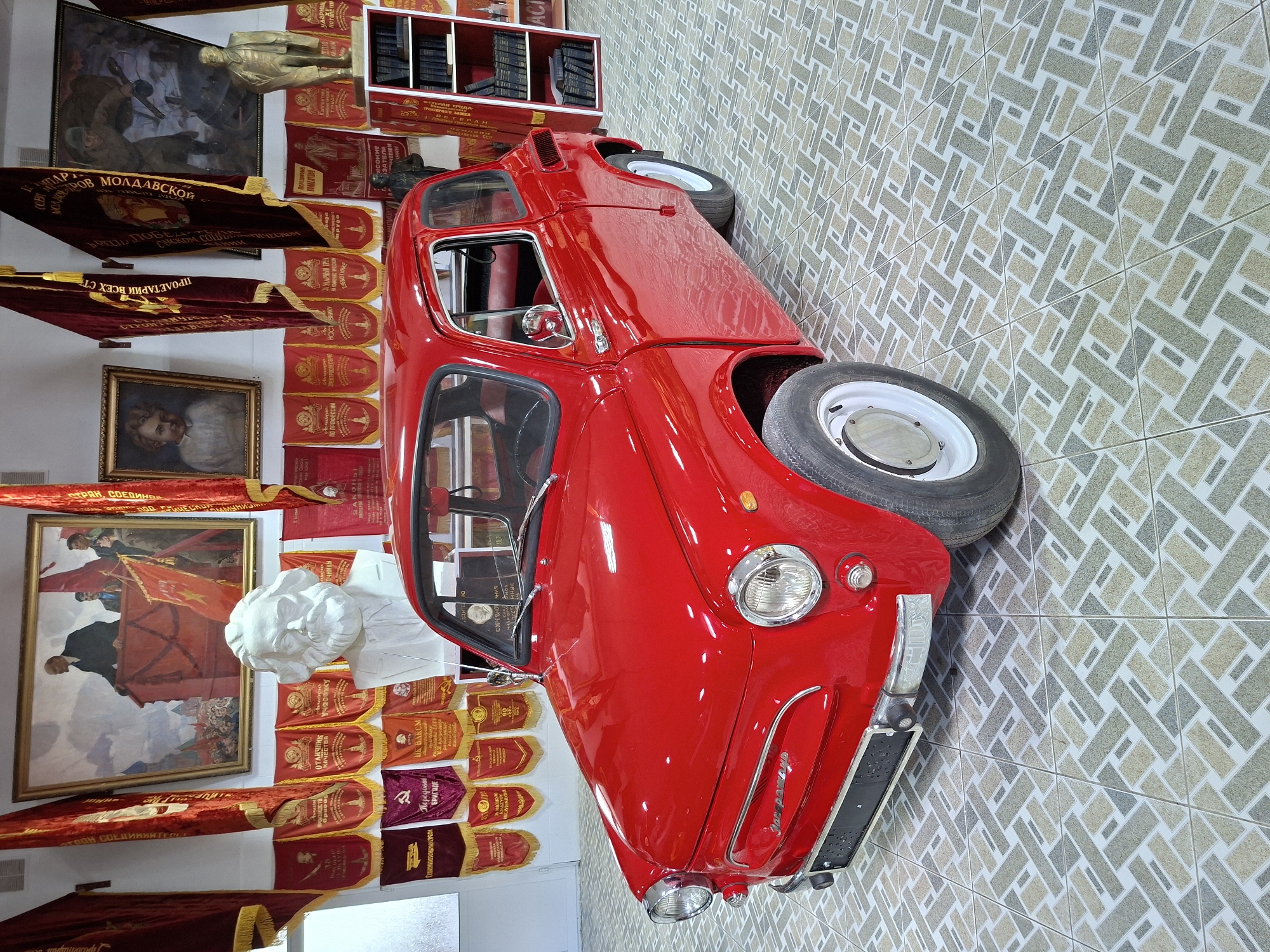 The image displays a red vintage car, likely a ZAZ-965 Zaporozhets, positioned in a room with a tiled floor. The car is the primary subject, facing left and slightly angled towards the viewer. Behind the car, the walls are adorned with a collection of Soviet-era propaganda materials, including paintings, flags, banners, and a bust of Karl Marx. To the right, a white bookshelf filled with dark-bound books is visible, alongside a red shelf containing more books. The overall setting suggests a museum or an exhibition space dedicated to Soviet history. While the image provides details about the objects within the scene, it does not offer specific information about people, actions, or interactions. The location of Ialoveni, Moldova, is mentioned, but the image itself does not contain visual cues to confirm or deny this specific location.