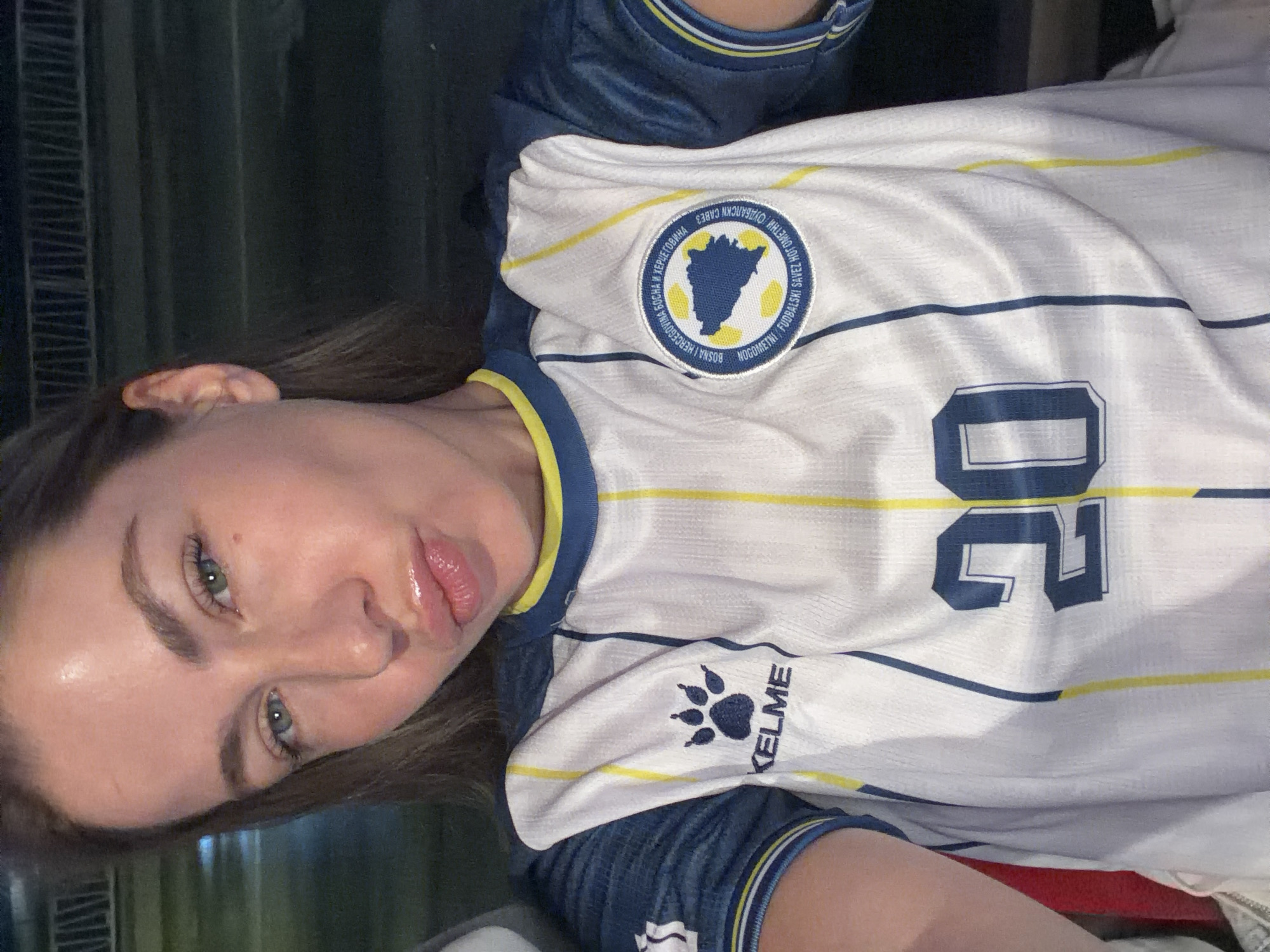 The image shows a close-up, selfie-style photograph of a young woman wearing a white and blue soccer jersey with yellow trim. The jersey has the number "20" prominently displayed on the front. A circular crest with a map of Bosnia and Herzegovina is visible on the left side of the jersey, along with text in Cyrillic and Latin scripts. The woman has her lips puckered, and her eyes are looking towards the camera. Her hair is brown and pulled back. The background is dark and out of focus, suggesting an indoor or nighttime setting, possibly a stadium or a dimly lit room. There are no discernible features in the background that would indicate a specific location within the city of Travnik, Bosnia and Herzegovina.