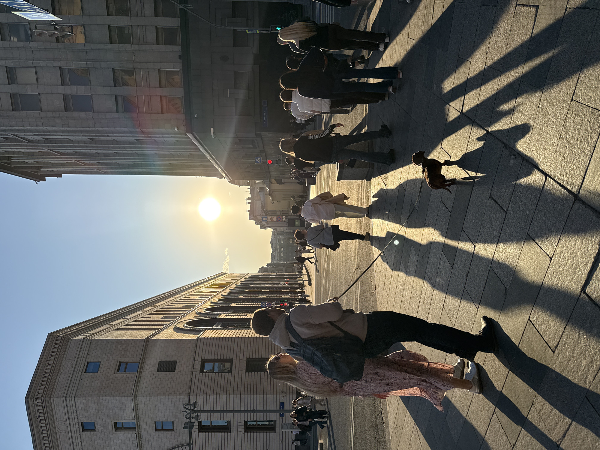 A long shot captures a street scene in Moscow, Russia. A couple stands in the foreground, a man with a backpack and a woman in a floral dress. They appear to be walking a small dog on a leash. Several other pedestrians are visible further down the sidewalk. The scene is illuminated by the setting sun, casting long shadows across the paved sidewalk. Buildings line both sides of the street, with architecture typical of central Moscow. Street signs in Russian are visible, and the overall setting conveys an urban environment at the end of the day.