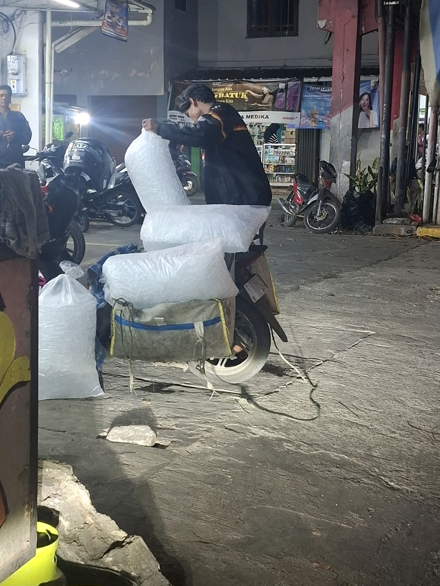 In Karangtengah, Indonesia, a person is loading a motorcycle with large plastic bags filled with ice. The motorcycle is parked on a paved street at night. The person is wearing a dark jacket and is in the process of securing the bags to the back of the motorcycle. Several other motorcycles are parked nearby, and a building with signage is visible in the background. The environment appears to be a commercial or street vendor area.