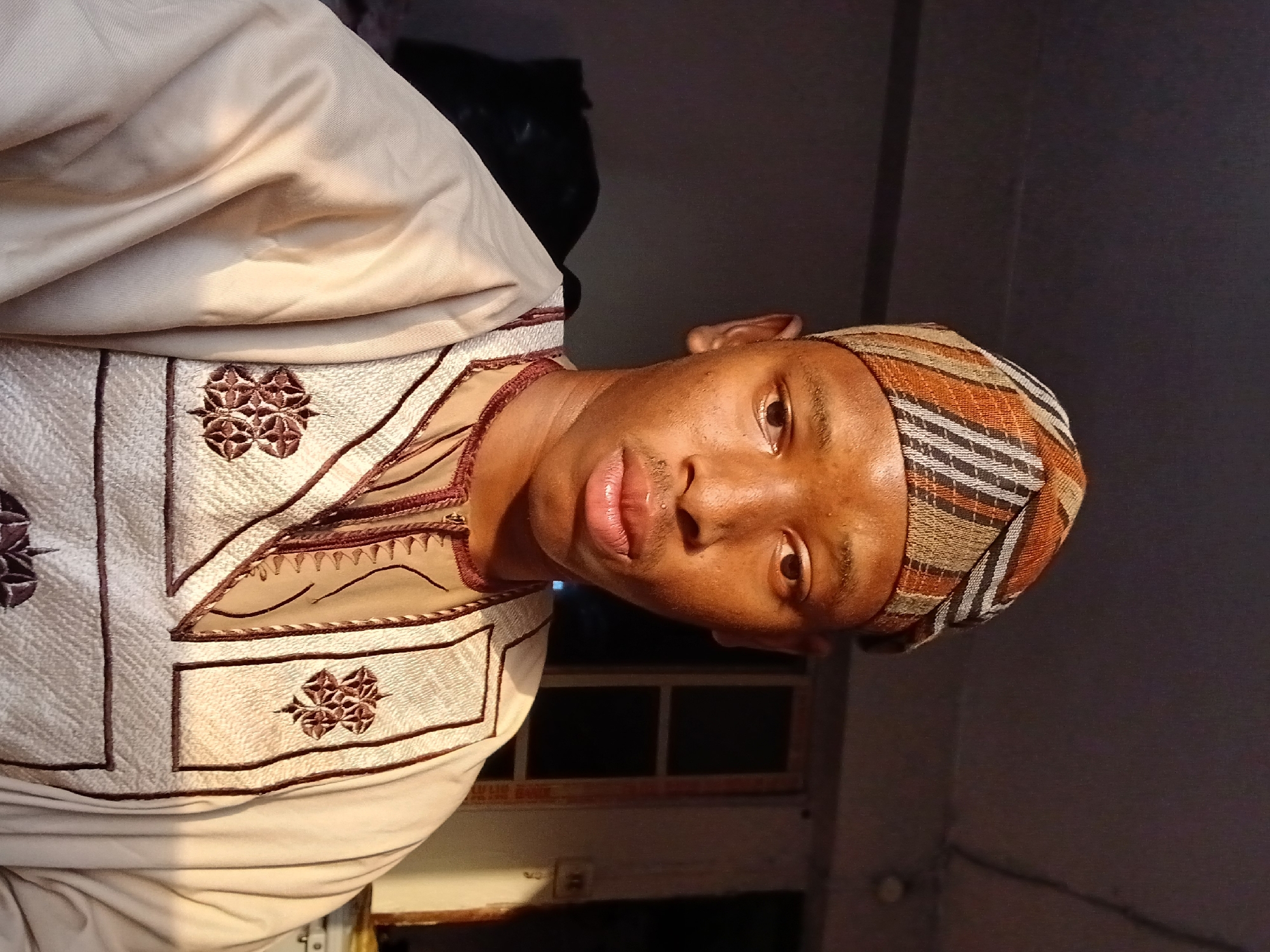 The image shows a man with dark skin, wearing a striped head covering and a light-colored garment with decorative embroidery. The background is indistinct but appears to be an interior space, possibly a room. There's insufficient information to establish the location in Lagos, Nigeria.