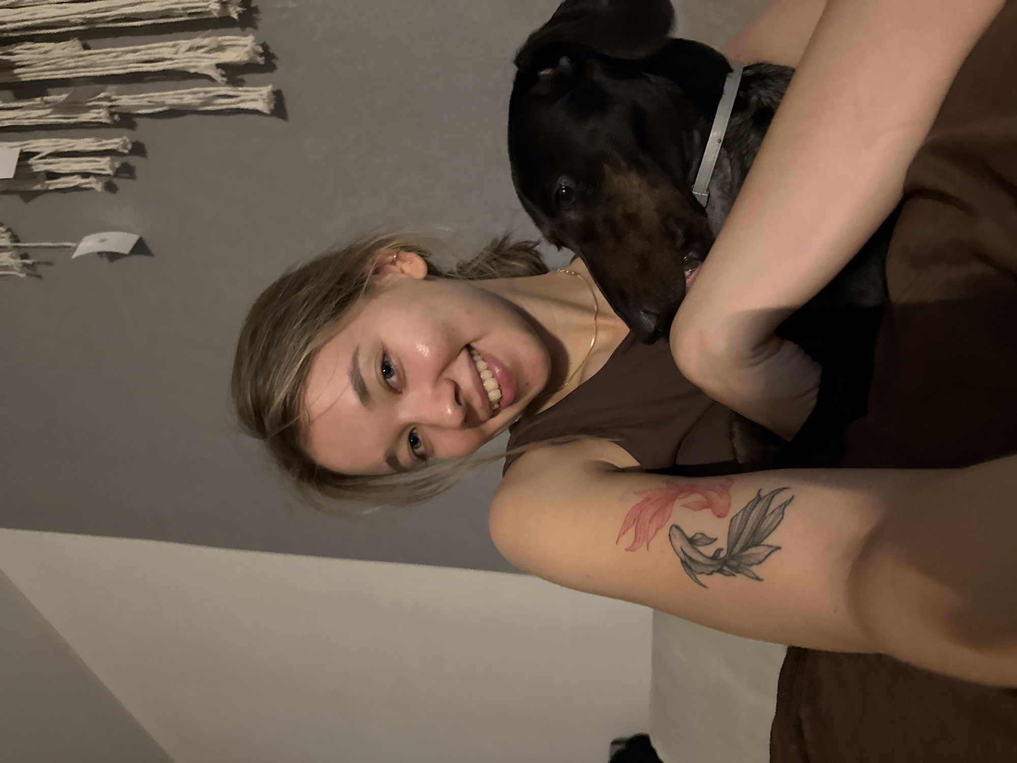 A young woman with blonde hair and blue eyes smiles at the camera, holding a black and brown dachshund. She has a tattoo of two koi fish on her left arm. The woman is wearing a brown tank top and a gold necklace. The dog is wearing a gray collar. They are sitting on a couch in front of a gray wall. A macrame wall hanging is visible on the wall behind them. The scene appears to be indoors, likely a living room. The location is not specified within Noviy, Russia.
