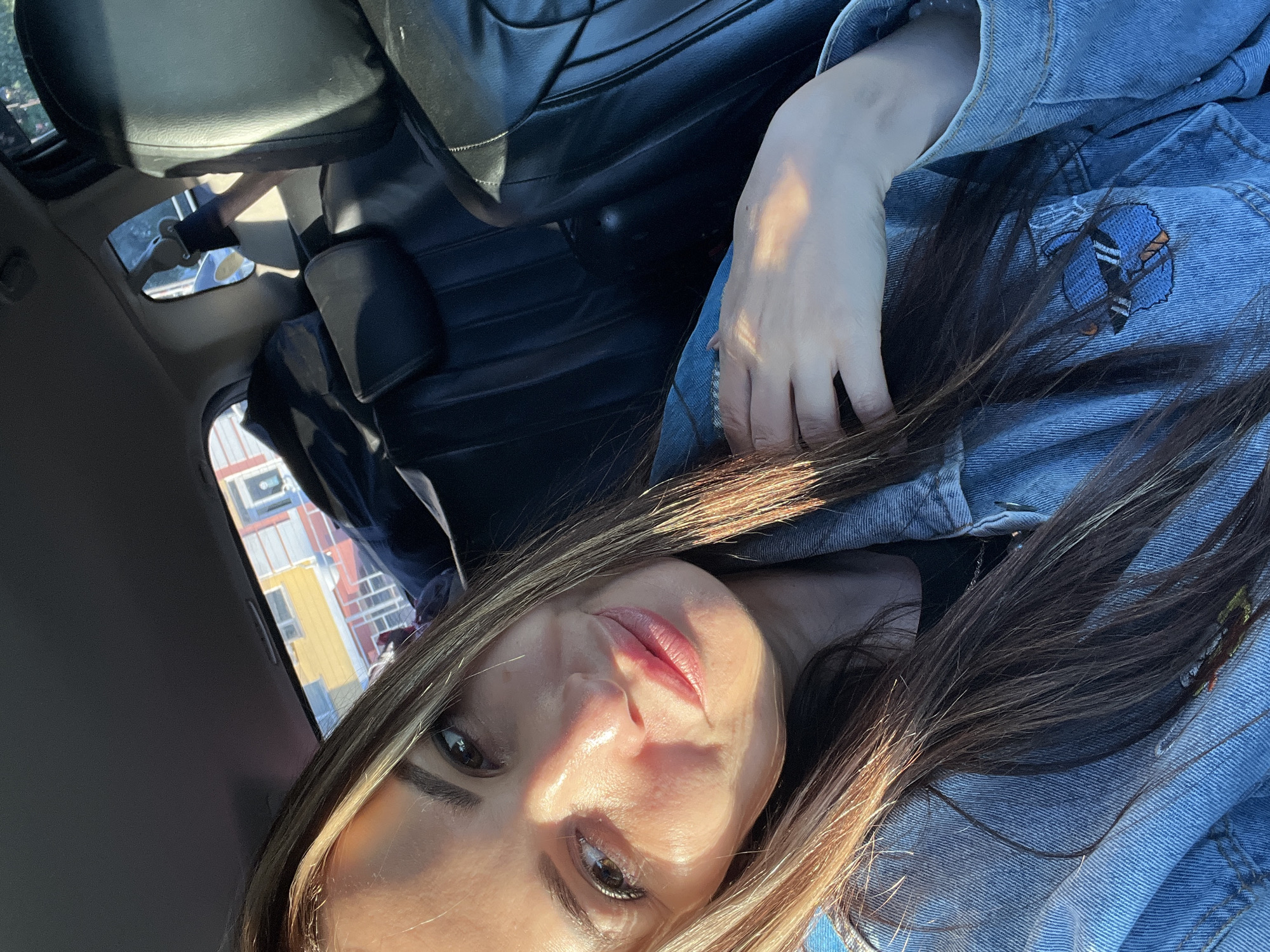 A woman is seated in a vehicle, possibly a car, with a partial view of a cityscape visible through the car window. The woman is wearing a denim jacket and has brown hair with blonde highlights. Her face is visible, and she appears to be looking at the camera. Part of the black leather interior of the car is also visible.