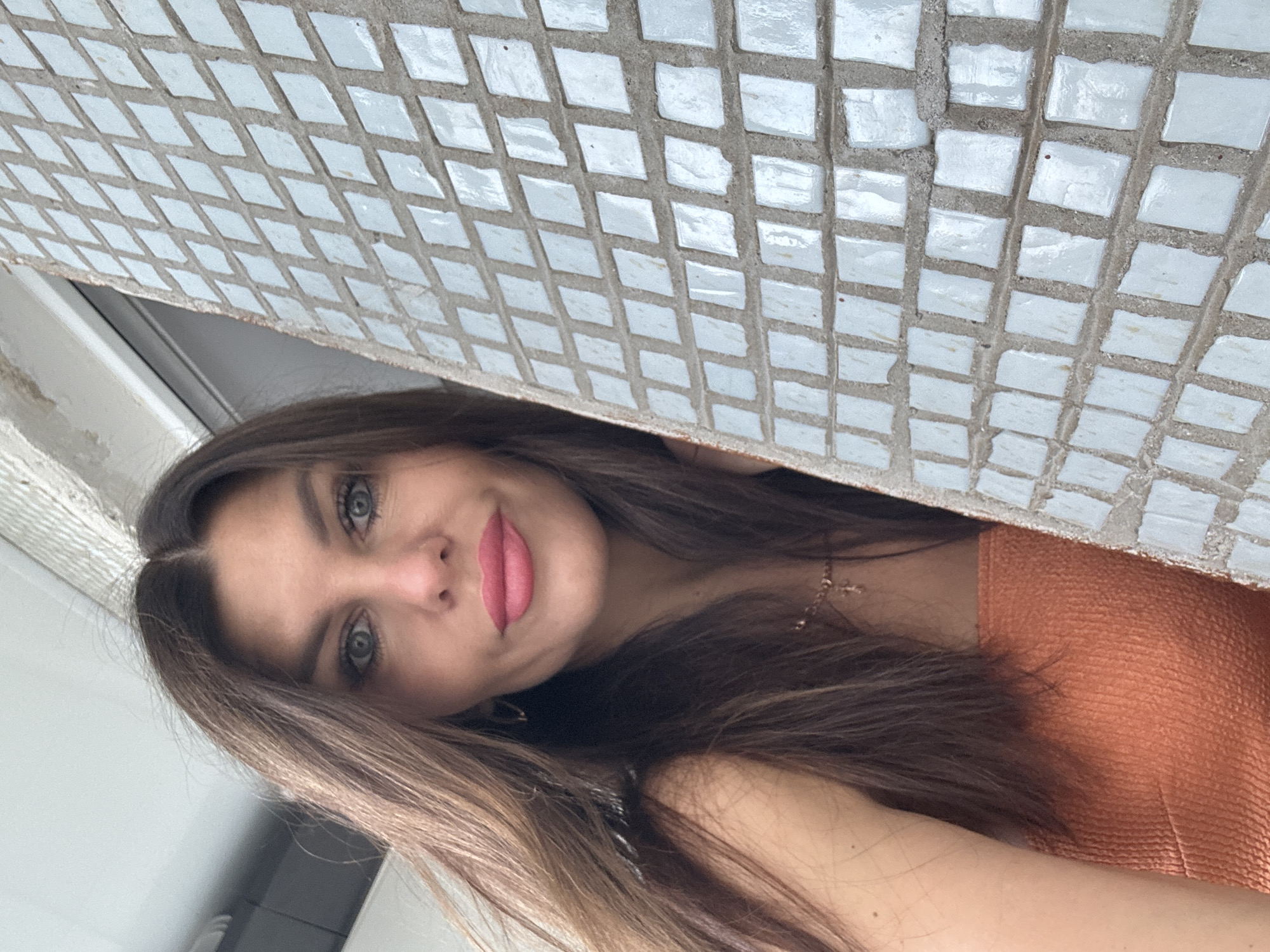 A woman with long, brown hair and blue eyes is pictured in a close-up shot, looking towards the camera. She is wearing a strapless orange top and a delicate gold necklace. To her right, a textured wall covered in small, square, light blue tiles is visible, creating a mosaic pattern. The background is slightly out of focus, showing a bright ceiling and a hint of a window frame. There are no discernible city landmarks in the image that would indicate a specific location within Chaikovsky, Russia.