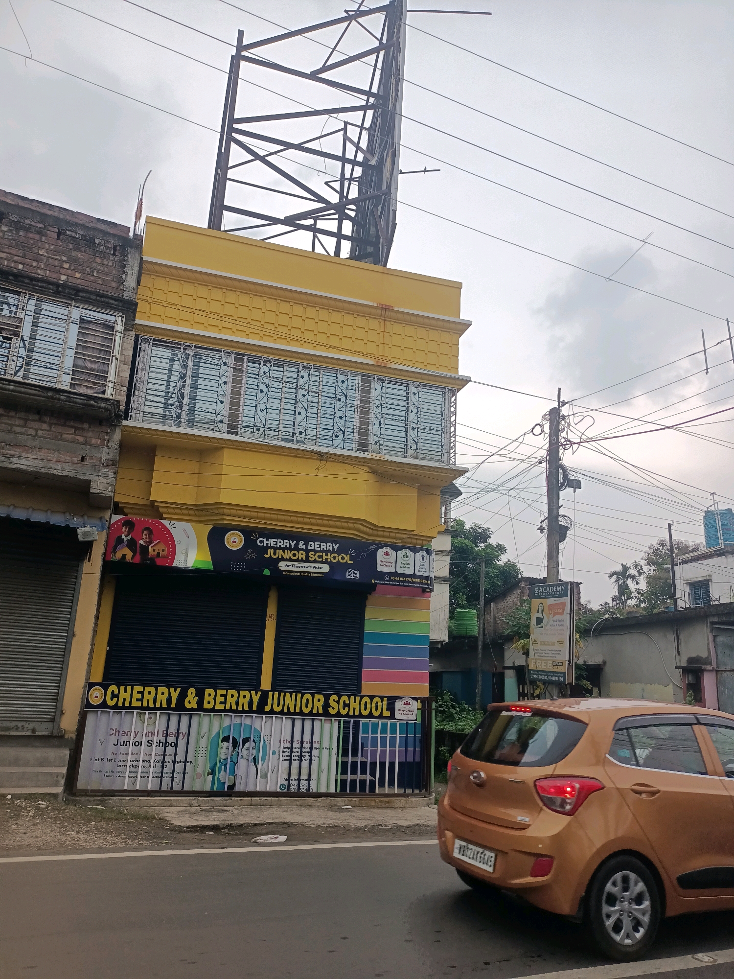 A street-level, eye-level, medium outdoor shot captures a yellow building with "CHERRY & BERRY JUNIOR SCHOOL" signage and a rainbow-colored mural on its facade. The building features metal-barred windows on the second floor and a dark, shuttered storefront on the ground floor. Adjacent to it, another building with brickwork and barred windows is visible. In the foreground, an orange Hyundai car, bearing a West Bengal license plate, is driving away from the camera. The background includes a utility pole with numerous tangled wires, trees, and other buildings. The sky is overcast. The scene appears to be in Barrackpore, India, as indicated by text on a sign for "The Academy" which also mentions "Barrackpore".