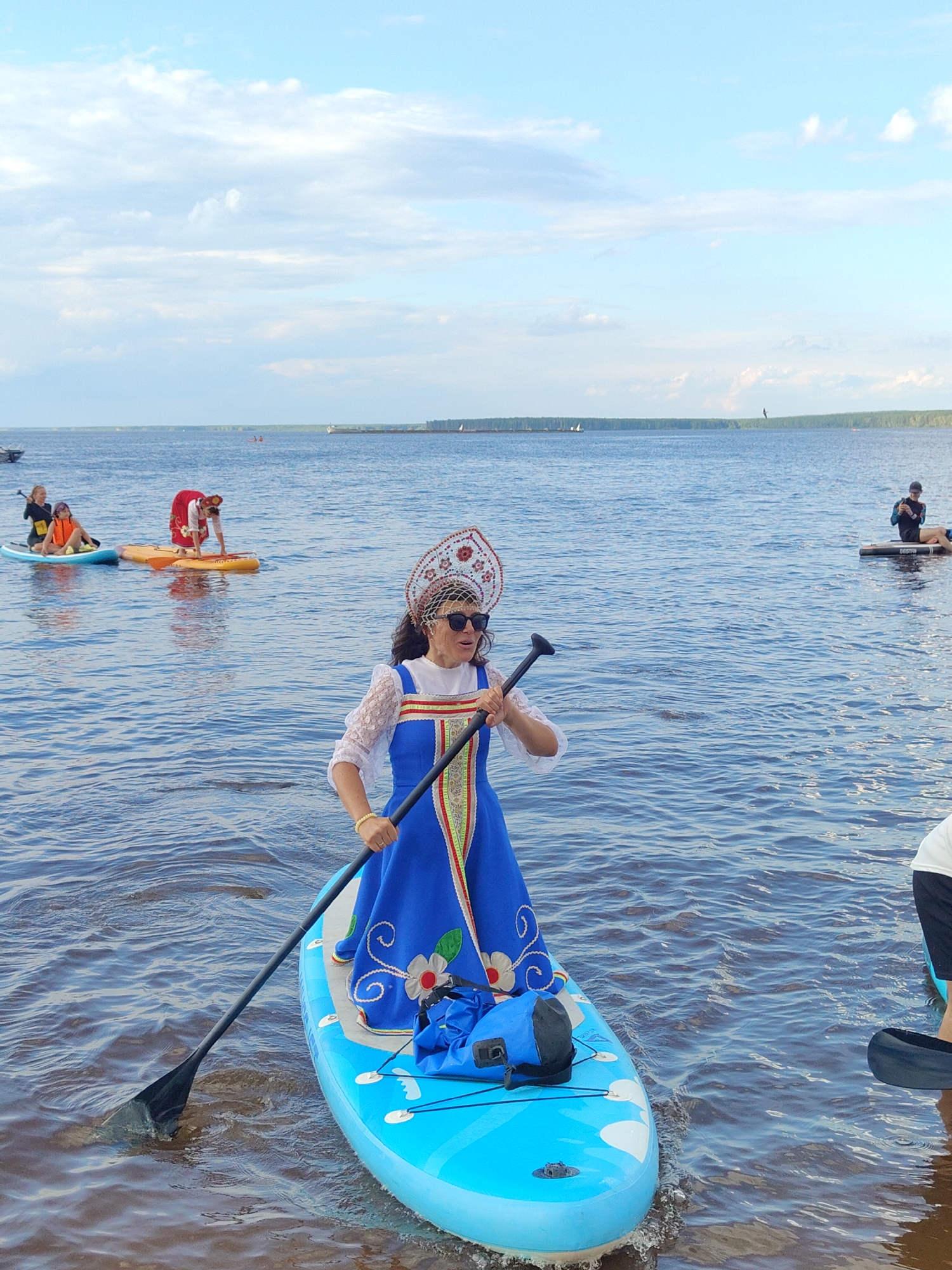 A woman in traditional Russian attire, including a kokoshnik headdress and a blue dress with red and white trim, is paddleboarding on a bright blue inflatable board in calm, dark blue water. She is holding a black paddle, with its blade submerged in the water. Sunglasses are on her face. In the background, two other paddleboarders are visible; one is on a yellow board and the other on a blue board. A person is sitting on a paddleboard further in the distance to the right, and another person is seen in the far distance to the right, also on a paddleboard. The sky above is a pale blue with scattered white clouds, and a treeline is visible on the horizon. The lighting suggests it is daytime.