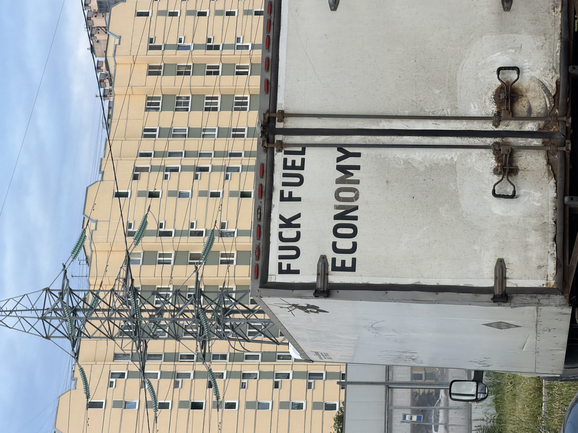 A white truck trailer is parked in front of a light-yellow building with many windows. The trailer has black text on its back doors that reads "FUCK FUEL ECONOMY". A large metal power line tower is visible in front of the building, with thick wires stretching across the frame. The sky is overcast and a green bush is visible on the left side of the frame.
