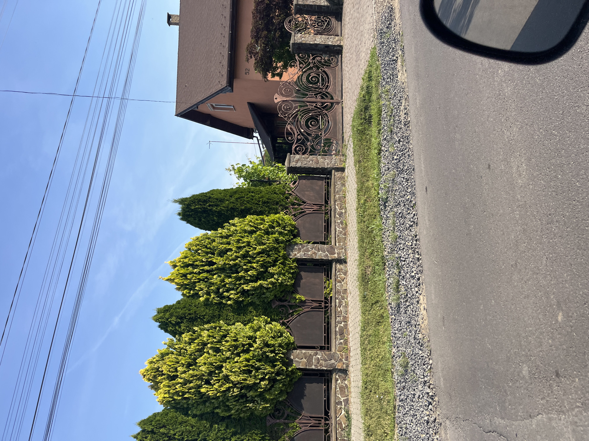 This image depicts a residential scene in Storozhnytsia, Ukraine. In the foreground, a grey asphalt road is visible, with a segment of a black car's side mirror in the bottom right corner. The road is bordered by a strip of grey gravel, followed by a narrow patch of green grass. Behind the grass is a decorative fence constructed with stone pillars and wrought iron panels, interspersed with closely planted evergreen trees, some of which have a yellowish-green hue. A large, ornate wrought iron gate is a prominent feature of the fence. Beyond the fence, a two-story house with a brown roof and reddish-orange walls is visible. The house has a chimney and a visible house number "52". The sky above is clear blue with a few faint white clouds. Electrical wires are strung across the upper portion of the image. The scene appears to be a sunny day.