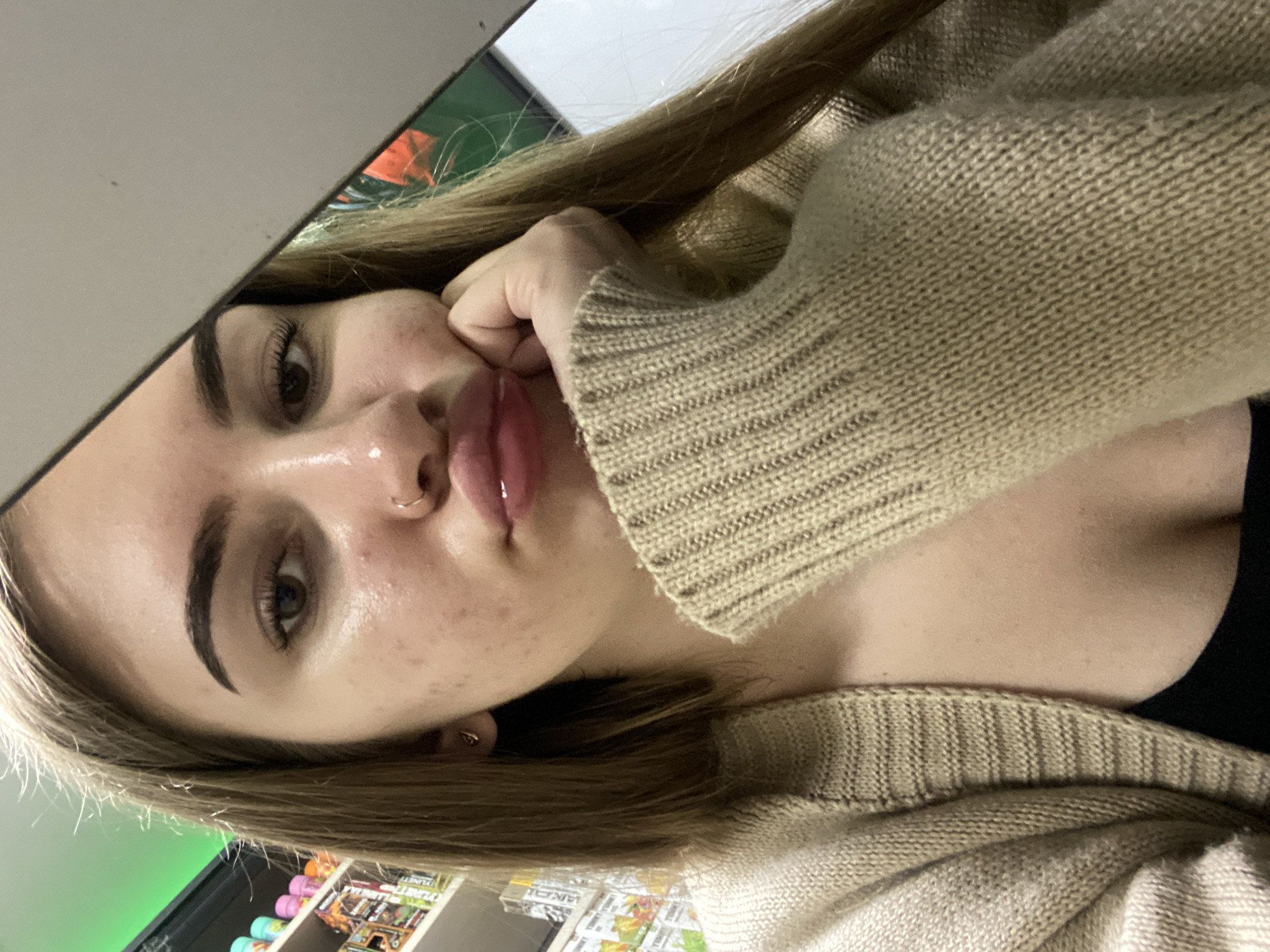 An overhead, tilted selfie captures a young person, likely female, from the chest up, looking directly at the camera. Her head is angled to the left, and her right hand rests against her cheek, partially obscuring her face. Her lips are slightly parted and appear glossy. She has long, light brown hair, prominent dark eyebrows, and dark eyes with visible eyelashes. A small hoop nose ring is visible on her right nostril. Her skin shows some texture, including visible pores and small marks. She is wearing a light beige or tan knitted sweater or cardigan with ribbed cuffs, and a dark garment is visible beneath it. The background is out of focus but appears to be an indoor setting, possibly a store. Shelves with various products, some brightly colored, are visible in the lower portion of the image. A section of a green sign or wall is discernible to the left. Faint text such as "PLANET" and "LAKAI" can be seen on product packaging in the bottom right corner. The lighting is artificial, consistent with an indoor environment. The overall impression is a casual, close-up portrait.