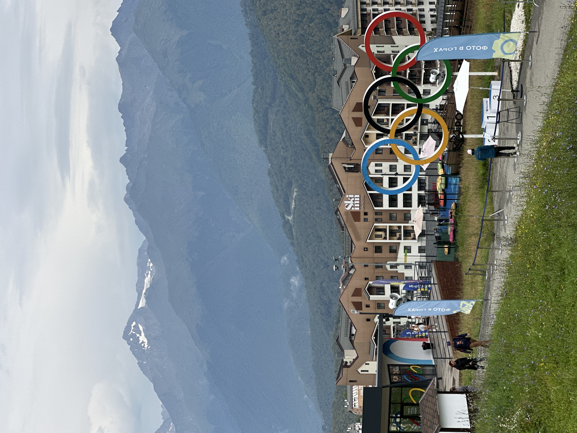 In the city of Unknown, Unknown, a group of people walk along a path in front of a hotel complex. The hotel is a multi-story building with a brown facade and numerous windows. In the foreground, a set of Olympic rings is displayed, with the blue ring on the left, followed by yellow, black, green, and red. A tall blue flag with text and an image of a camera stands to the right of the Olympic rings. The path is bordered by grass and wildflowers. In the background, a mountain range with snow-capped peaks rises under a cloudy sky. The weather appears to be overcast.