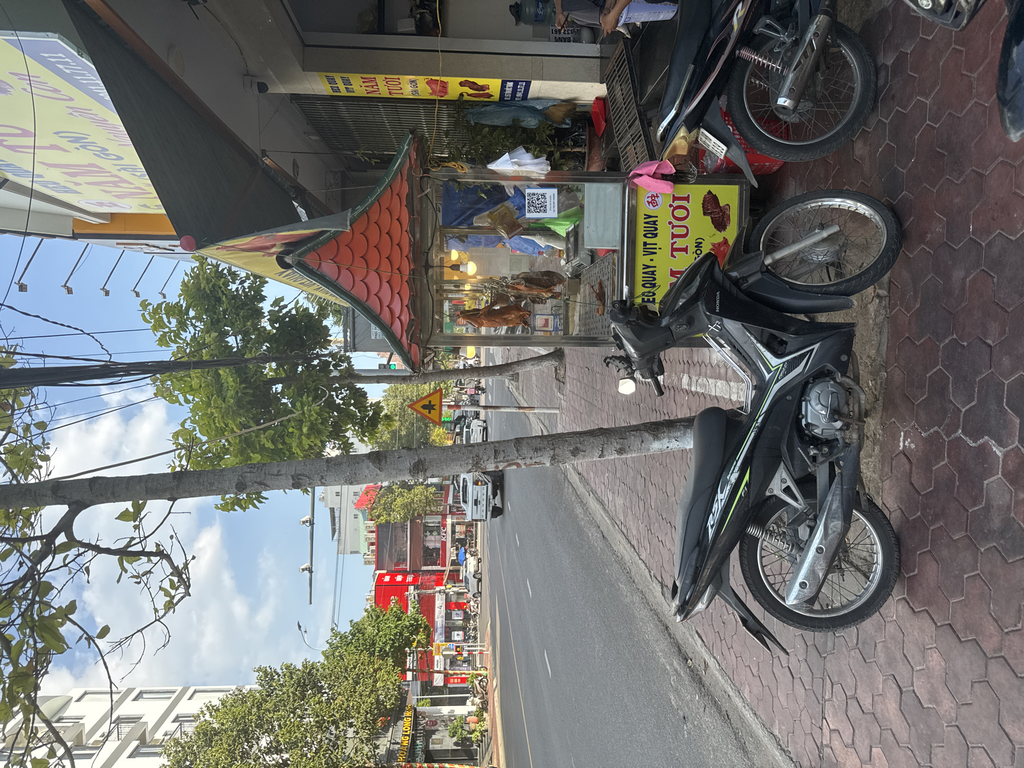 The image shows a street scene in Phú Thủy, Vietnam. A sidewalk is paved with red hexagonal bricks, and a tree with a textured trunk stands near the sidewalk. A black motorcycle is parked on the sidewalk, facing right. Further down the sidewalk, a small food stall with a red, tiled roof displays several roasted ducks hanging inside a glass enclosure. A yellow sign with Vietnamese text and images of food is attached to the stall. Next to the stall, another motorcycle is partially visible. Across the street, there is a multi-lane road with traffic, including cars and motorcycles. Several buildings line the street, some with bright red facades. The sky is partly cloudy.