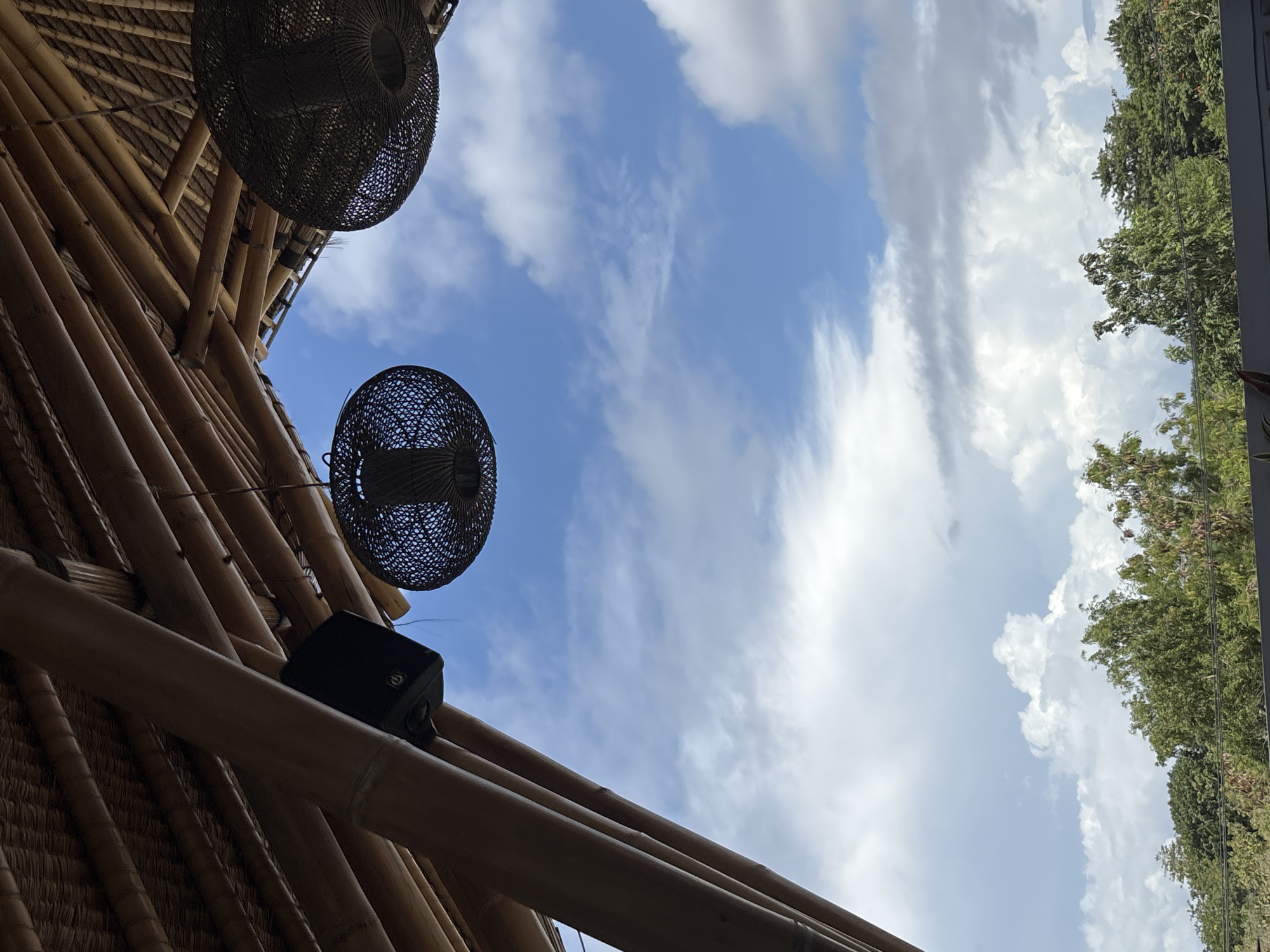 The image, taken from a low angle, shows a section of a bamboo structure against a backdrop of a partly cloudy sky. Two woven, globe-shaped light fixtures hang from the structure. A small, rectangular black object is attached to the bamboo. Trees are visible in the upper right corner of the frame. The image suggests an outdoor space, likely a building's exterior or an open-air structure. The context of Kuta Selatan, Indonesia, indicates a tropical environment.
