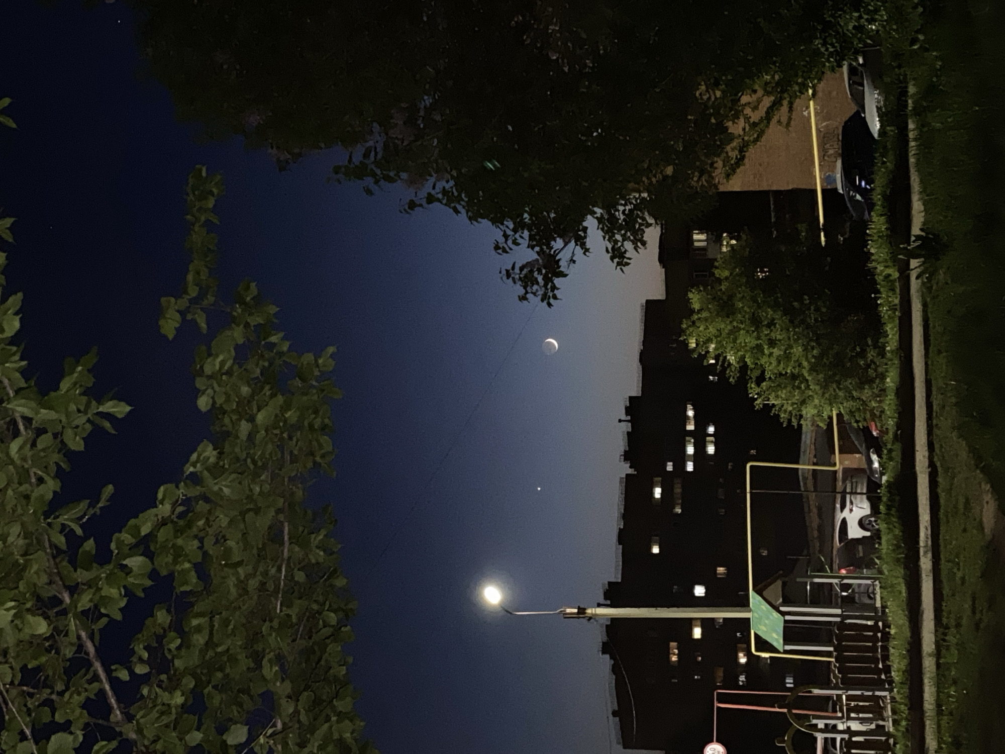The image presents an urban scene at night, likely in Votkinsk, Russia. In the center of the frame is the crescent moon in the night sky above a street light, and a smaller star. Building silhouetted with illuminated windows are present. Trees frame the sky. In the lower right quadrant, is a playground. Adjacent to the playground, a sidewalk has a car parked on the curb.