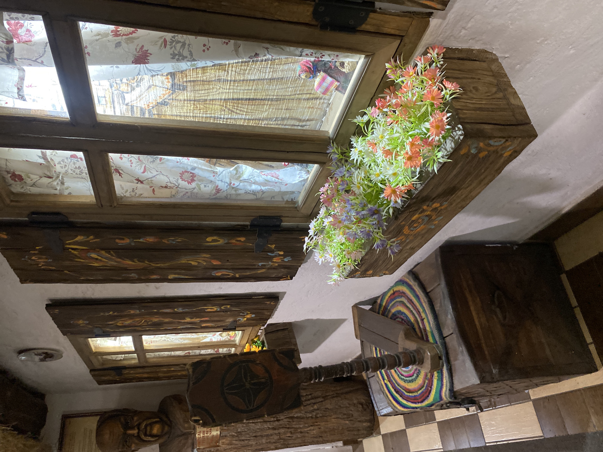 The image shows the interior of a room with rustic decor, possibly in a restaurant or guesthouse. There are two windows visible, both framed with wood and appearing to look out onto another building or street. The windows have floral-patterned fabric serving as curtains or shades. A wooden planter with artificial flowers is attached to the wall near the upper window. The walls are white. Below, a checkerboard floor pattern is visible.
A wooden chair is nearby, as well as a carved wood sculpture.
Location information given as Noviy, Russia.
