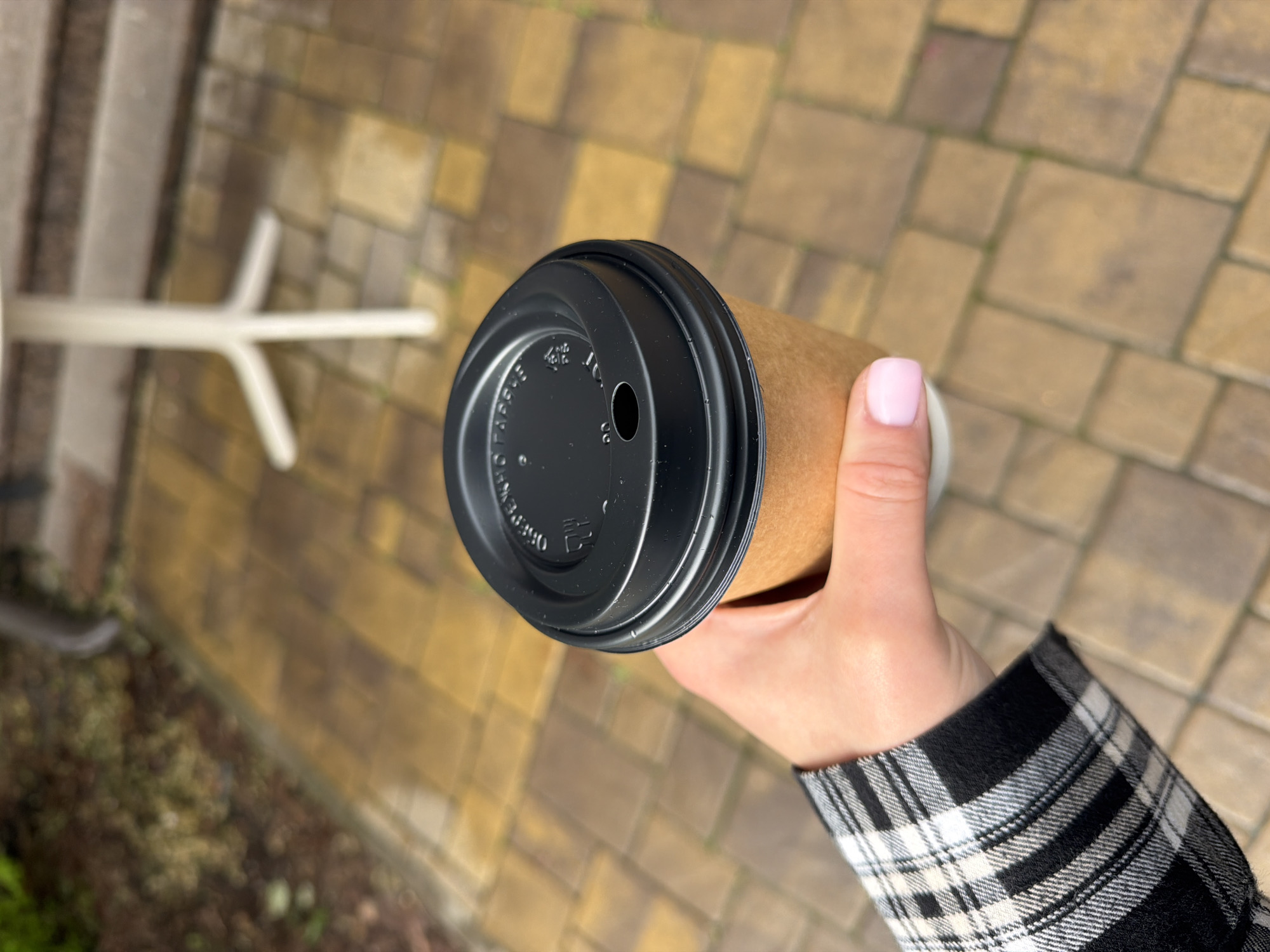 The image presents a close-up, high-angle view of a person's hand holding a brown paper coffee cup in an outdoor setting. The hand, which has light pink nail polish on the thumb, is gripping the cup. The person is wearing a black and white plaid sleeve, suggesting a casual shirt or jacket.
The coffee cup is a standard kraft paper type, fitted with a black plastic lid. On the lid, white text is visible around the edge. The phrase "KEEP CALM AND DRINK COFFEE" can be discerned, and the word "HOT" is faintly visible near the circular sipping hole.
The background consists of an outdoor paved area, featuring rectangular pavers in various shades of light brown, tan, and grayish-brown, with traces of green growth in the cracks. To the left, a blurry white, Y-shaped object, possibly a base for outdoor furniture or a table leg, is partially visible. Further down and to the left, blurred dark green foliage or ground cover can be seen.
The scene occurs during the daytime, evidenced by the natural light and shadows, indicating a bright or sunny day. There are no discernible emotions, portraying a casual, everyday moment. This activity of holding a coffee cup likely takes place while walking or standing in an outdoor area in Sofiivska Borshchahivka, Ukraine.