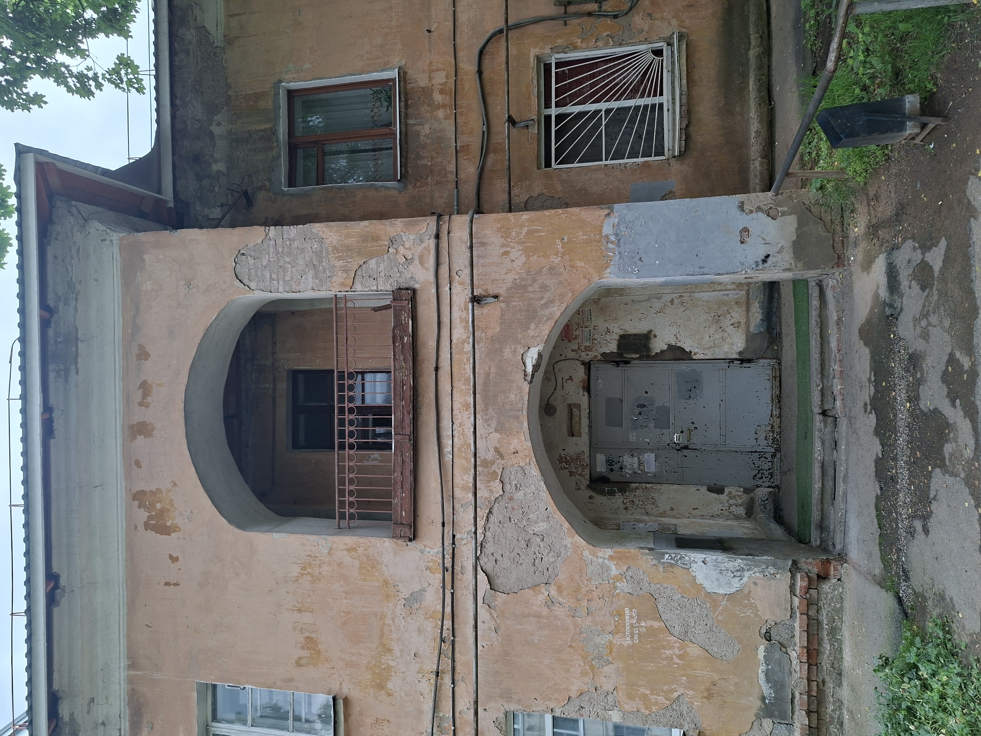 The image shows the facade of an old, dilapidated apartment building in Izhevsk, Russia. The building is made of brick and stucco, with peeling paint and visible signs of decay, such as cracked plaster and exposed brickwork. There are two arched entrances on the ground floor, one of which has a metal door with a lock. Above the ground floor, there is a balcony with a metal railing. The windows of the building are old-fashioned, with wooden frames, and some are covered with protective bars. Electrical wires are visible running along the facade. The ground in front of the building is paved with concrete and asphalt, with patches of grass and weeds growing through. A metal fence and a trash can are visible to the right of the building. The sky is overcast, suggesting cloudy weather. The building appears to be residential, likely an older apartment block.