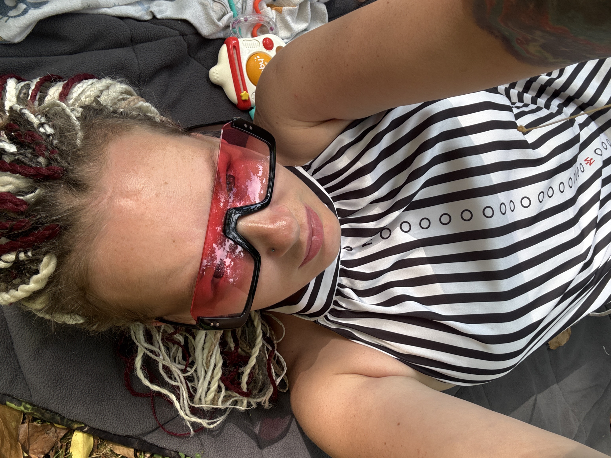 The media file features a close-up, overhead shot of a person lying down outdoors. The subject has a nose ring and is wearing large, translucent pink sunglasses. Their hair is styled in thick braids, with some strands in a reddish-brown color and others in a blonde or white hue. They are wearing a black and white striped garment, possibly a dress or top, with a pattern of small black circles and some red text on the right side of the garment. To the upper right of the subject, a portion of a child's toy is visible, appearing to be a red and white electronic toy with a yellow circular button.
The scene is set outdoors, as indicated by the presence of fallen leaves and the dappled sunlight on the subject's arm and the garment. The ground appears to be covered with a dark gray blanket or mat, with some lighter-colored fabric or clothing also visible. The lighting suggests it is daytime, likely sunny. The overall mood appears relaxed and casual.
There is visible red text on the striped garment, appearing to read "SE" followed by a series of circles. The image context places this scene in Sochi, Russia.