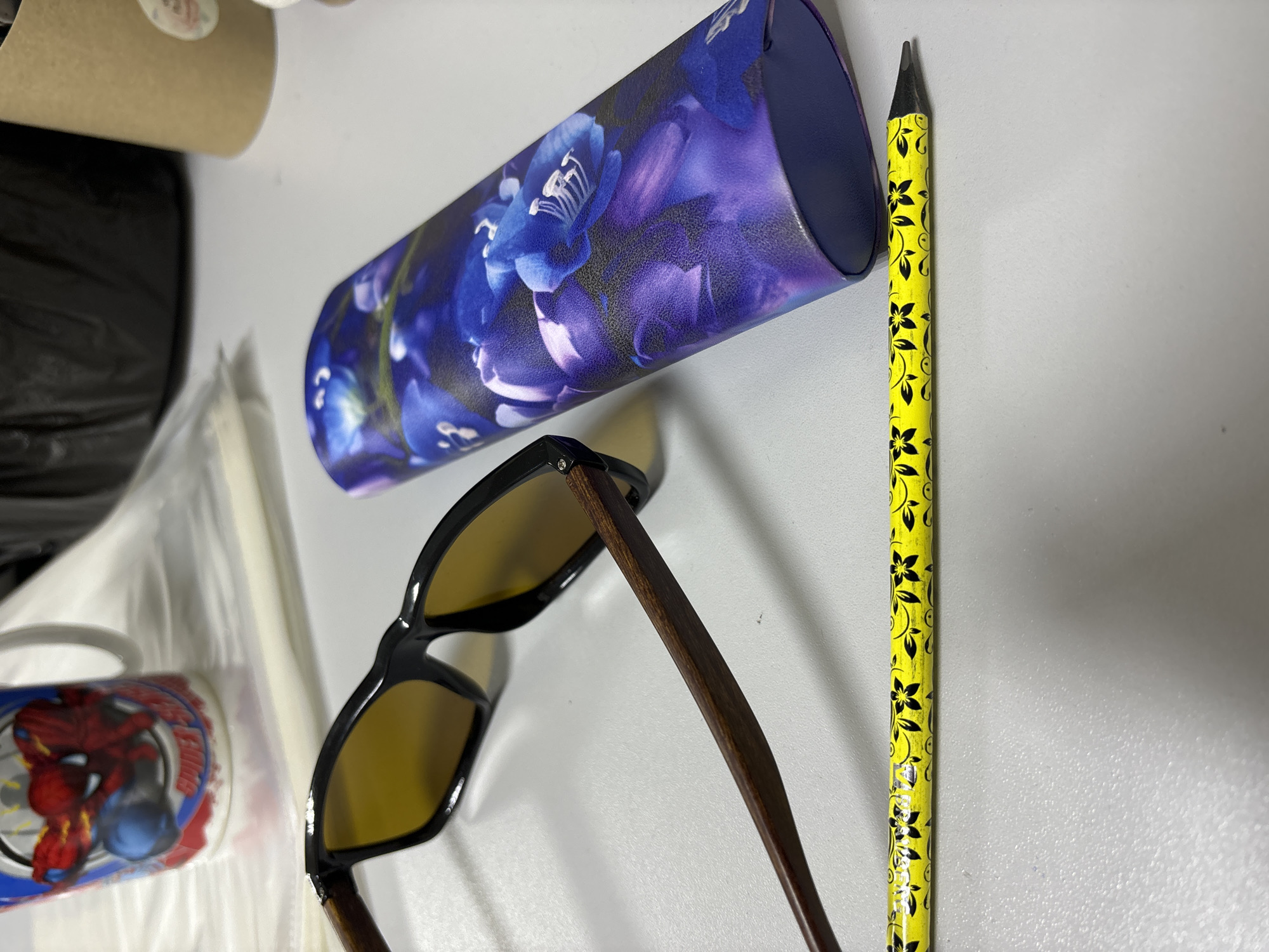 The scene presents an eye-level, indoor shot. There are multiple objects laid on a white surface, including a pair of sunglasses, a pencil, and a floral case. The sunglasses have a black frame and wooden temples with amber lenses. Adjacent to the sunglasses, there is a yellow pencil adorned with a black floral pattern. Beside the sunglasses is a cylindrical case decorated with a blue floral design. Part of a mug with a Spider-Man design is visible in the left side of the frame.
The setting of Chaikovsky, Russia, cannot be definitively determined from the given visual information.