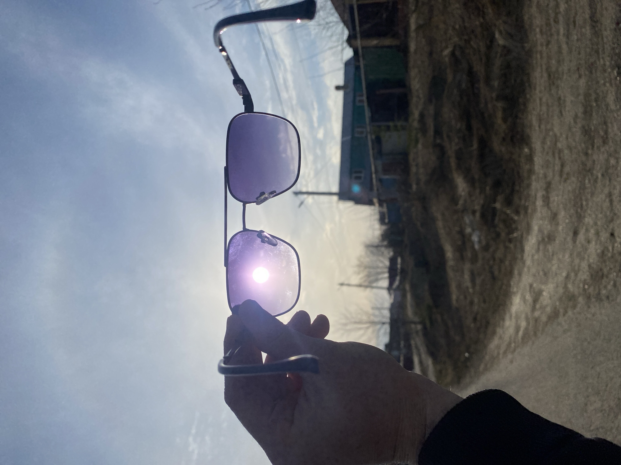 A hand holds up a pair of aviator sunglasses against a bright sky with some clouds. The sun is visible through the lower lens. In the blurred background is a street scene with buildings and trees, likely in an urban area. The road is paved with a dark-colored material. The location is Kazan, Russia.