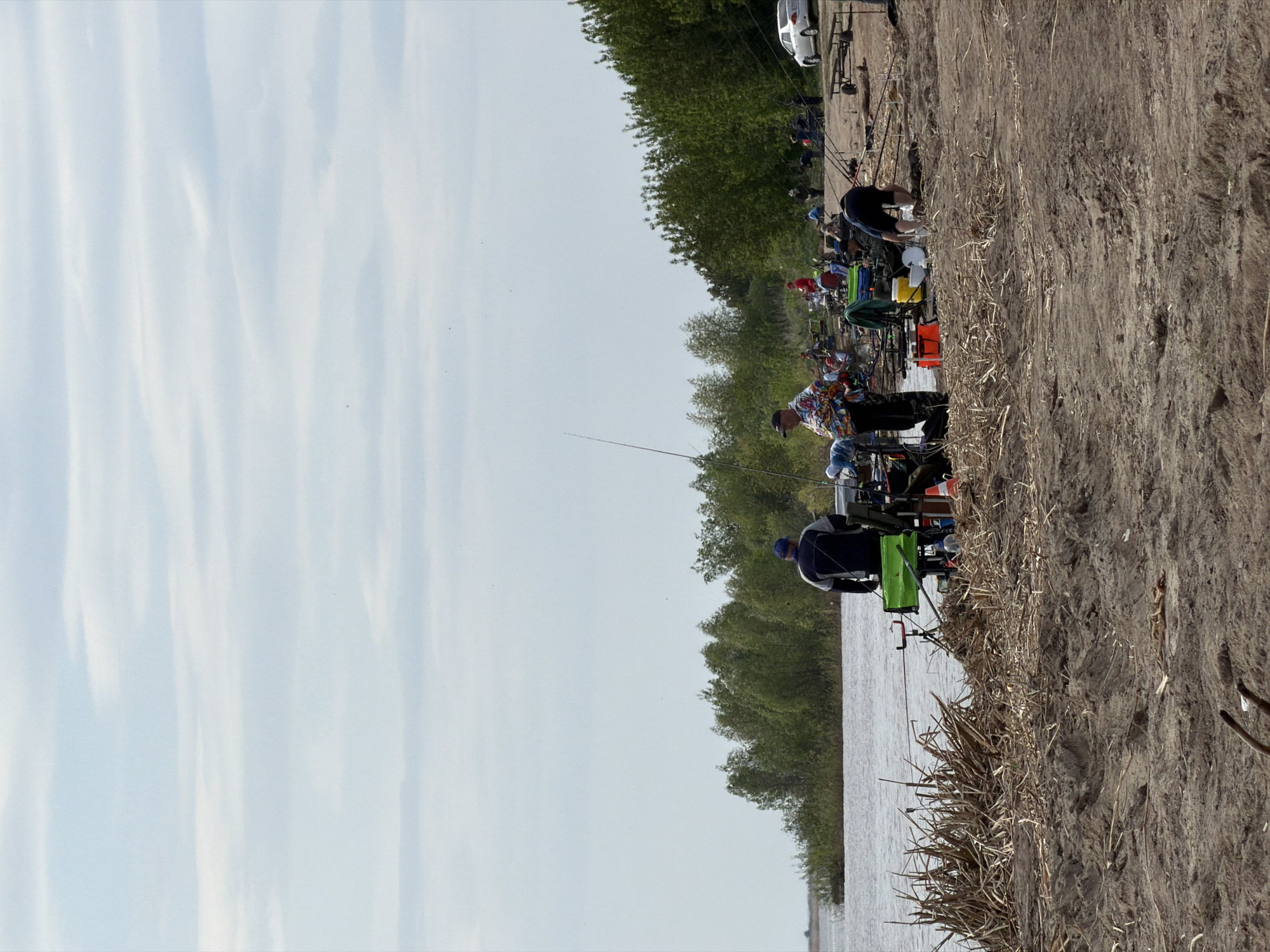 The image shows a group of people fishing along a riverbank in what appears to be Krasnyi Bor, Russia. Multiple individuals are spaced along the shore, each with fishing rods extended towards the water. Fishing equipment such as chairs, tackle boxes, and rod holders are visible at each station. The river bank is composed of earth and dry vegetation. Trees line the opposite side of the river. The sky is overcast. A vehicle and trailer can be seen in the background.