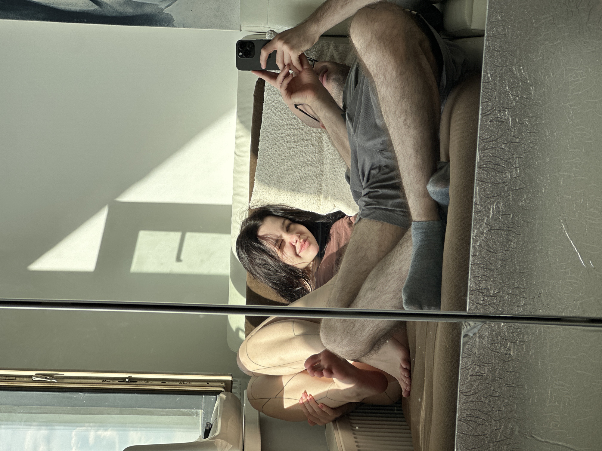 The image appears to be a selfie taken in a mirror. A man is holding a smartphone in his right hand, pointed towards the mirror. He is wearing glasses and a grey t-shirt. He is seated next to a woman on a couch. The woman has dark hair and is smiling slightly. The couch is beige. There is a window with sunlight shining through it in the background. The location is Khimki, Russia.
