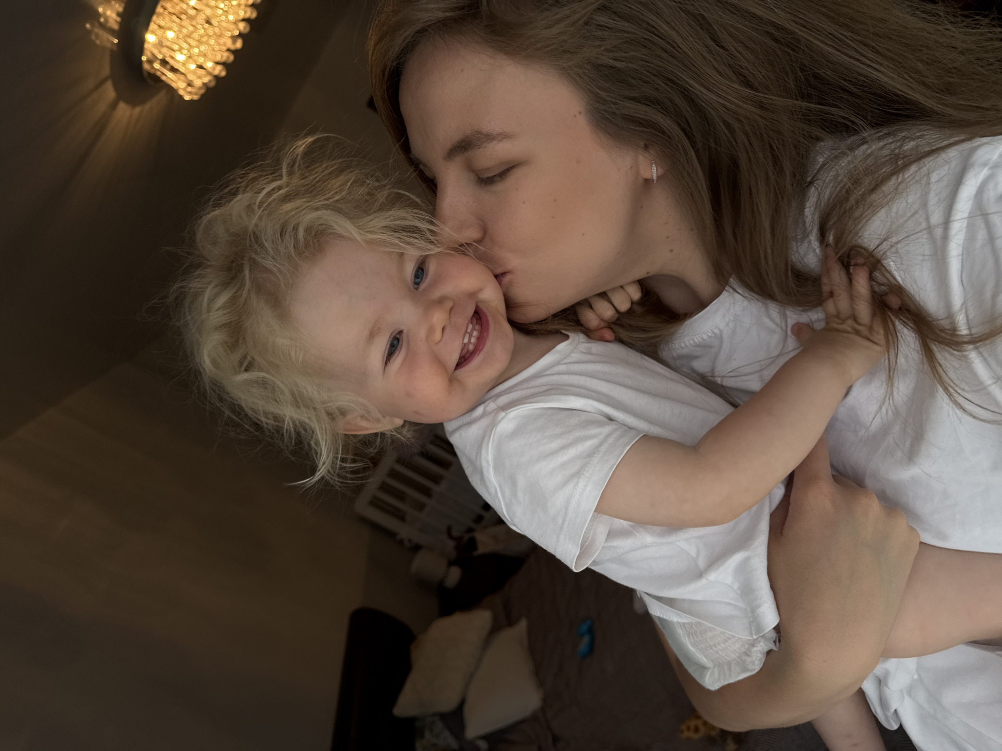 In this close-up, candid shot, a woman with long, brown hair is kissing a young child on the cheek. Both are wearing white t-shirts. The child, with blonde, curly hair and blue eyes, is looking towards the camera with a wide, joyful smile, revealing a gap between their front teeth. The woman's eyes are closed as she leans in for the kiss.
The scene appears to be indoors, likely a bedroom. A dark brown, textured surface, possibly a bed or rug, is visible in the background, along with white pillows. To the upper left, a chandelier with multiple sparkling lights casts a warm glow and creates dramatic shadows on the ceiling, suggesting it's evening or the room is dimly lit. The overall mood is one of affection and tenderness. The location context indicates this is in Vladimir, Russia. No visible text is present in the image.