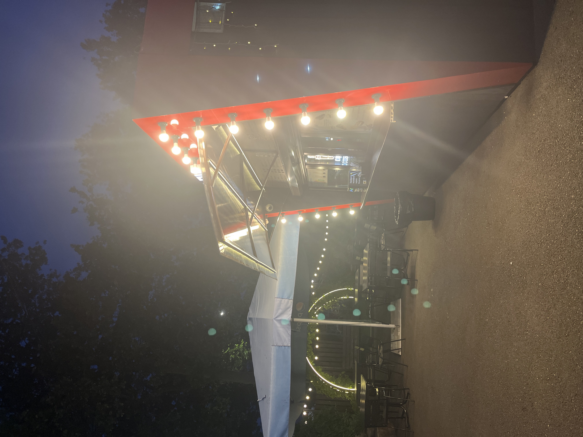The image depicts a food stand at night, likely in an outdoor seating area. The food stand itself is modern, with a black exterior and a prominent red, angular accent. It is adorned with a string of bright, exposed light bulbs, suggesting a lively atmosphere. To the left, a large, light blue umbrella shades a seating area with tables and chairs. String lights are draped around the umbrella and further into the background, creating a festive ambiance. A circular archway, also illuminated, stands behind the tables. The ground appears to be paved. The sky above is a deep twilight blue, with a faint glow from what might be the moon visible through the trees in the background. The overall scene suggests a casual dining or gathering spot. There are no people visible in the image. The image has been taken in Krasnodar, Russia.