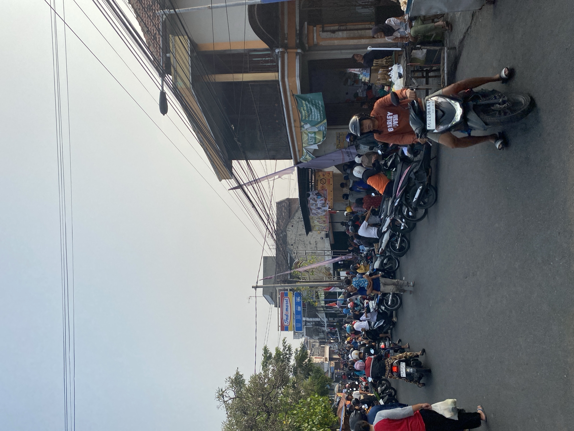 A street scene in Kediri City, Indonesia, displays a busy atmosphere with numerous motorcycles and pedestrians. The image captures a road lined with buildings and shops, suggesting a commercial or residential area. Many people are on or around motorcycles, indicating transportation and daily life activities. Overhead, electrical wires are visible, common in urban Indonesian settings. One building has a prominent sign for "Indomaret," a popular convenience store chain in Indonesia, which helps to identify the location. A man in an orange hoodie is riding a motorcycle in the foreground, his face partially obscured by a helmet. The overall scene conveys a sense of bustling activity and community engagement on a street in Indonesia.