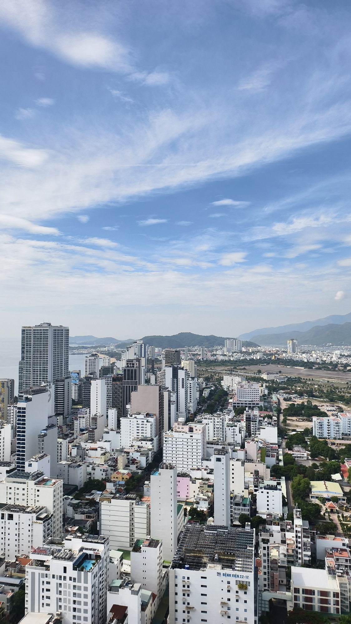 This is an aerial, full-frame, vertical, eye-level shot of a dense urban cityscape in Nha Trang, Vietnam, under a partly cloudy blue sky. The foreground and midground are filled with numerous high-rise buildings of varying heights and architectural styles, predominantly white and grey, with some featuring balconies, windows, and rooftop amenities like swimming pools. A few smaller, colorful residential buildings are interspersed among the skyscrapers. In the background, more buildings extend towards rolling green hills and mountains that form a backdrop to the city, with a hint of the ocean visible on the left. The overall impression is one of a bustling coastal city with extensive development.