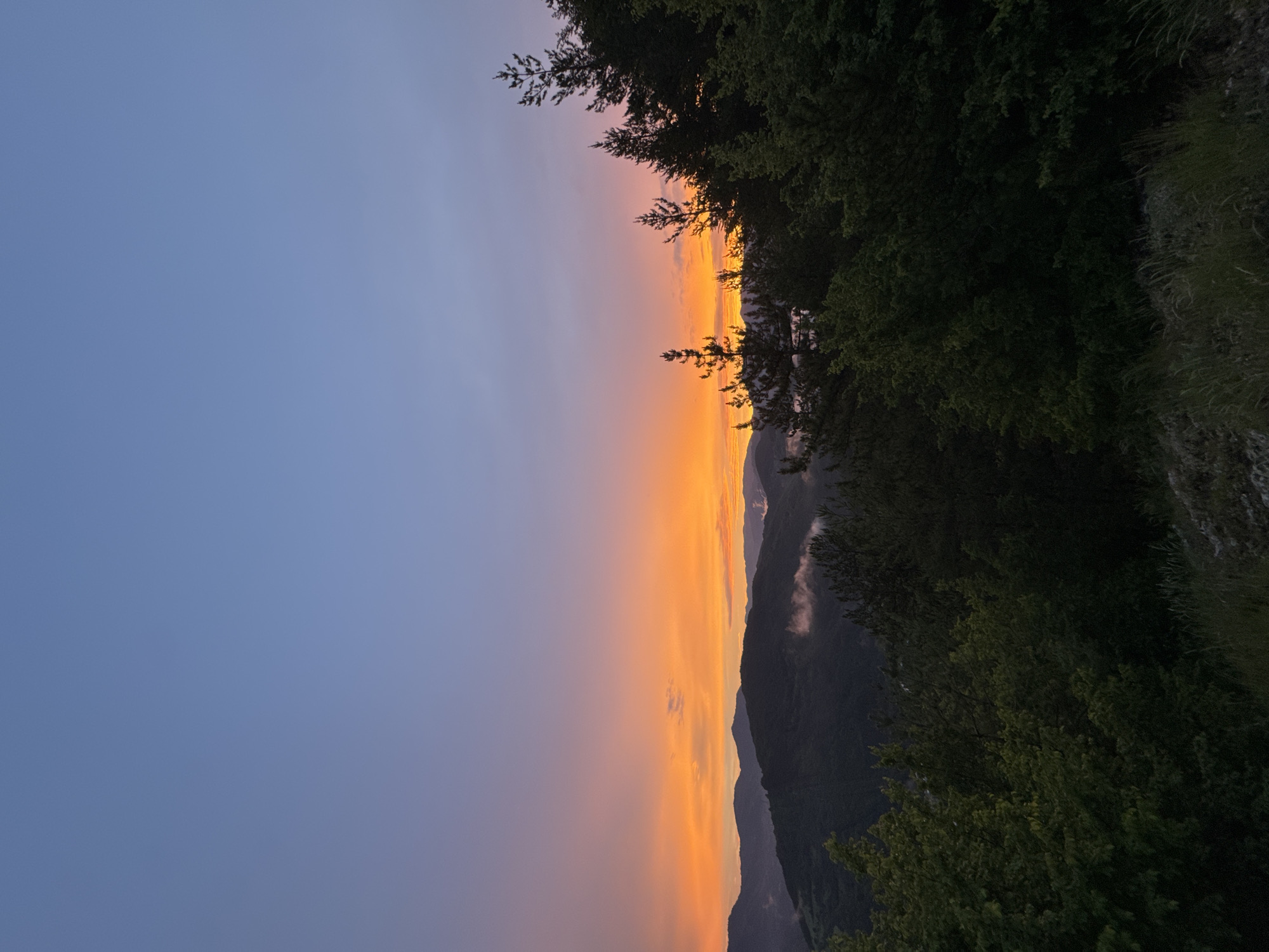 The image depicts a mountainous landscape at dusk. The sky is a gradient of soft blue at the top, transitioning to hues of pink and vibrant orange near the horizon, indicating sunset or sunrise. Layered, dark green mountains recede into the distance, with some patches of white mist clinging to their slopes. In the foreground, a dense forest of evergreen and deciduous trees is visible, silhouetted against the colorful sky. The trees are lush and green, with some taller pine trees reaching upwards. The overall scene is serene and natural, evoking a sense of peace and the beauty of the wilderness. The location is the city of Strazha, Bulgaria.