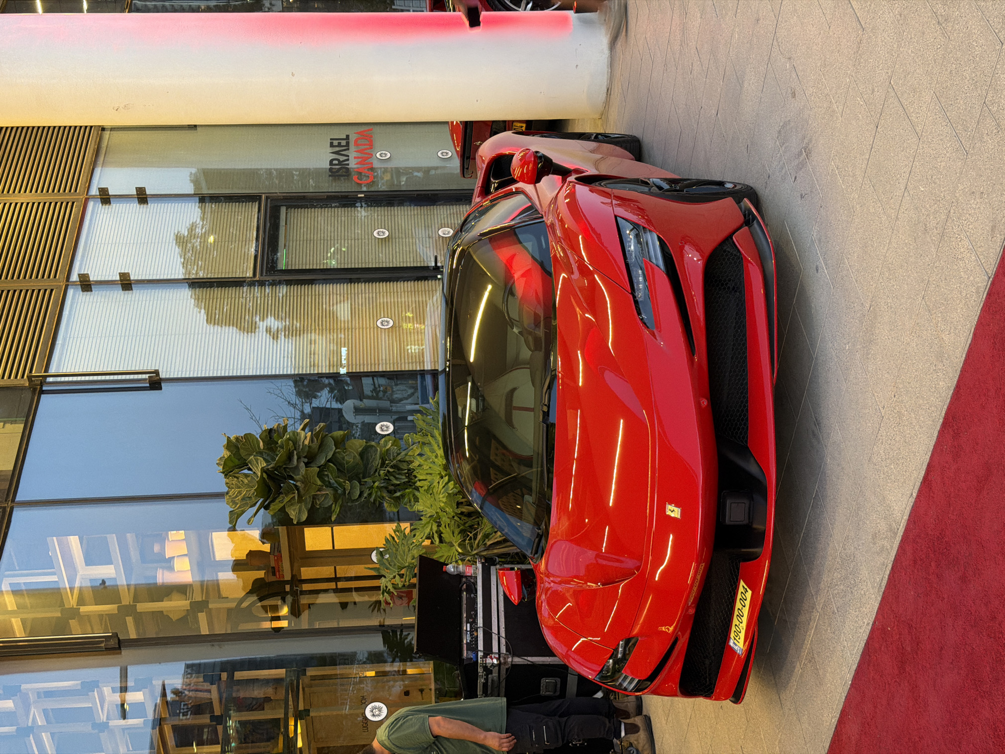 A red Ferrari with Israeli license plates is parked in front of a building in Tel Aviv, Israel. The building has a glass facade that reflects the surroundings. A red carpet is visible on the ground next to the car. There is a person standing near the front of the car, and sound equipment is positioned on the ground behind it. "Israel Canada" is visible on the building's facade.