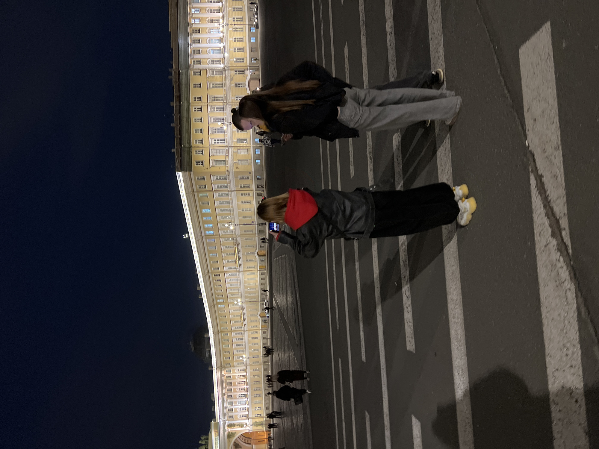 Two young women stand side-by-side in Palace Square, Saint Petersburg, Russia, illuminated by the building behind them and photographing its lighted façade. The building appears to be the Winter Palace. Pedestrians are seen in the background, near the palace arches. The street surface is marked with painted crosswalk lines. One woman wears a black jacket and grey pants. The second woman wears a black jacket with a red hoodie.