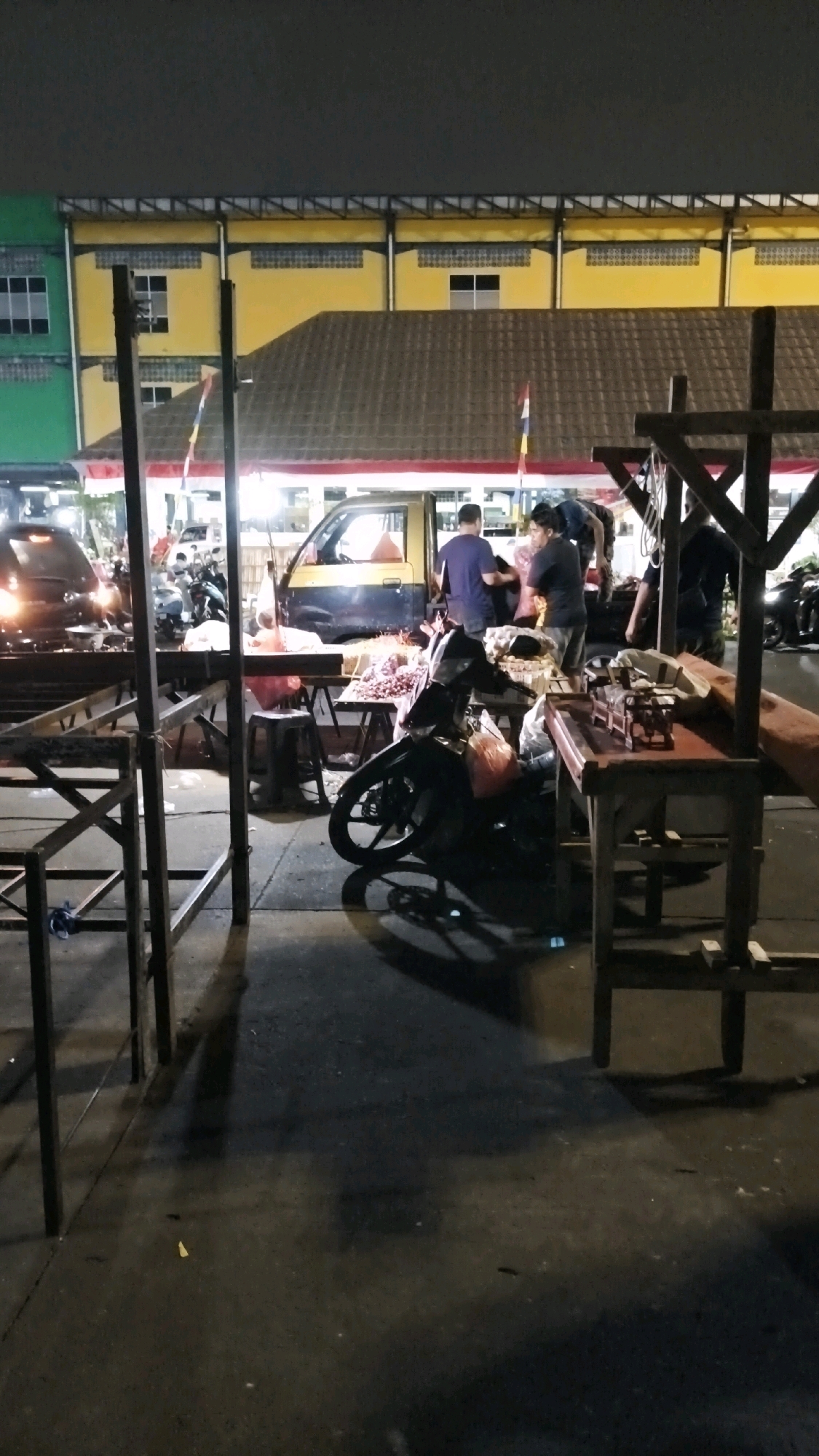 The image depicts an outdoor market scene at night in South Jakarta, Indonesia. In the foreground, a metallic market stall structure with wooden planks is visible. A motorcycle is parked in the middle ground, with a pile of produce, likely onions, on a table next to it. Several people are gathered around another stall where a yellow and blue van is parked. They appear to be engaged in activity related to the market, possibly selling or buying goods. In the background, a building with green and yellow sections is illuminated, with what appears to be a tiled roof. The ground is wet, reflecting the ambient light. The overall scene suggests a bustling night market environment.
