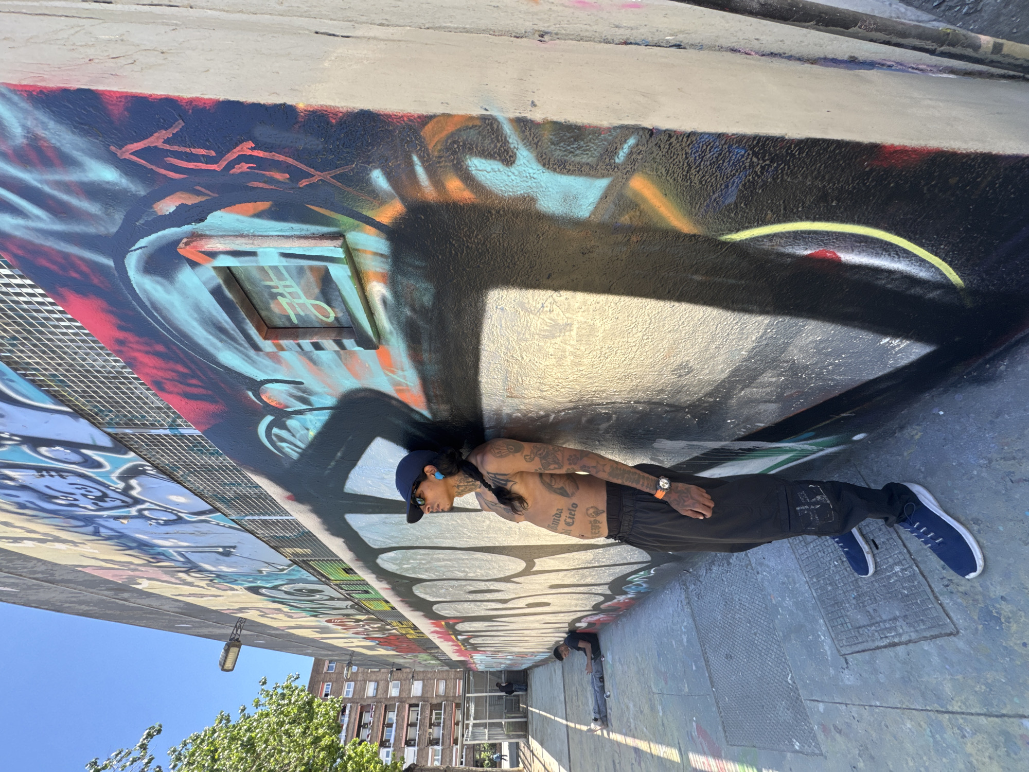 A man with tattoos is leaning against a graffiti-covered wall under a bridge or overpass. He is wearing a blue cap, sunglasses, a bare chest, black pants, and blue sneakers. He also has a watch on his left wrist. The graffiti on the wall is colorful and abstract.
In the background, other people are visible, and there is also greenery, buildings, and a clear blue sky, which indicates an outdoor setting. The image is rotated, making the ground appear to be on the side.
Without explicit location markers within the image, definitively confirming it is Barcelona requires additional context.