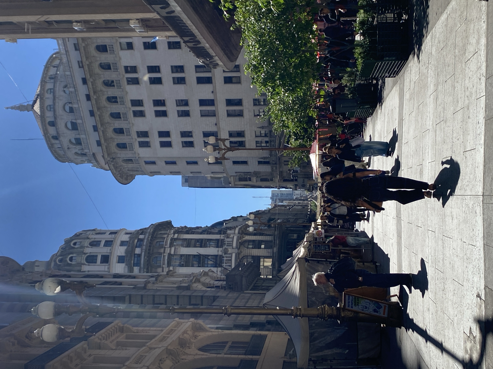 This image captures a bustling, sunlit pedestrian street in Buenos Aires, Argentina, characterized by its grand architecture and lively atmosphere. The scene is dominated by two prominent classical-style buildings under a clear blue sky. On the left, a distinctive multi-tiered building features an ornate dome-like roof with numerous arched windows, strongly resembling the iconic Palacio Barolo. Another large, multi-story building, also with classical elements and rectangular windows, lines the right side of the street.
The wide, paved walkway is filled with people, indicating a busy urban environment. In the foreground, a man with white hair, wearing a dark jacket and trousers, stands next to a display board. The board exhibits Spanish text, "PROTECTORES DE PANTALLA" (Screen Protectors), and a cartoon graphic. His long shadow, along with others stretching across the light-colored pavement, suggests strong sunlight, likely in the morning or late afternoon.
Further down the street, more people are visible, some gathered under a canopy of green trees. Several white awnings or market stalls are present, hinting at an outdoor market or vendors. Ornate streetlights punctuate the scene, and green park benches are visible under the trees. The weather appears bright and clear, contributing to the vibrant energy of the street.