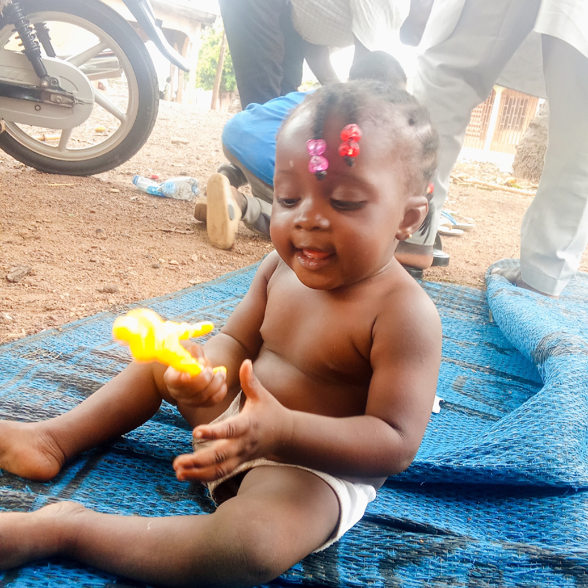 The image shows a baby sitting on a blue woven mat in what appears to be an outdoor location in Akwanga, Nigeria. The baby is dark-skinned, wearing a white diaper, and has colorful hair decorations on their forehead. The baby is holding a yellow toy in their hands. In the background are other people, a parked motorcycle, and a water bottle on the ground. The ground is dirt. A building is partially visible behind the people.
