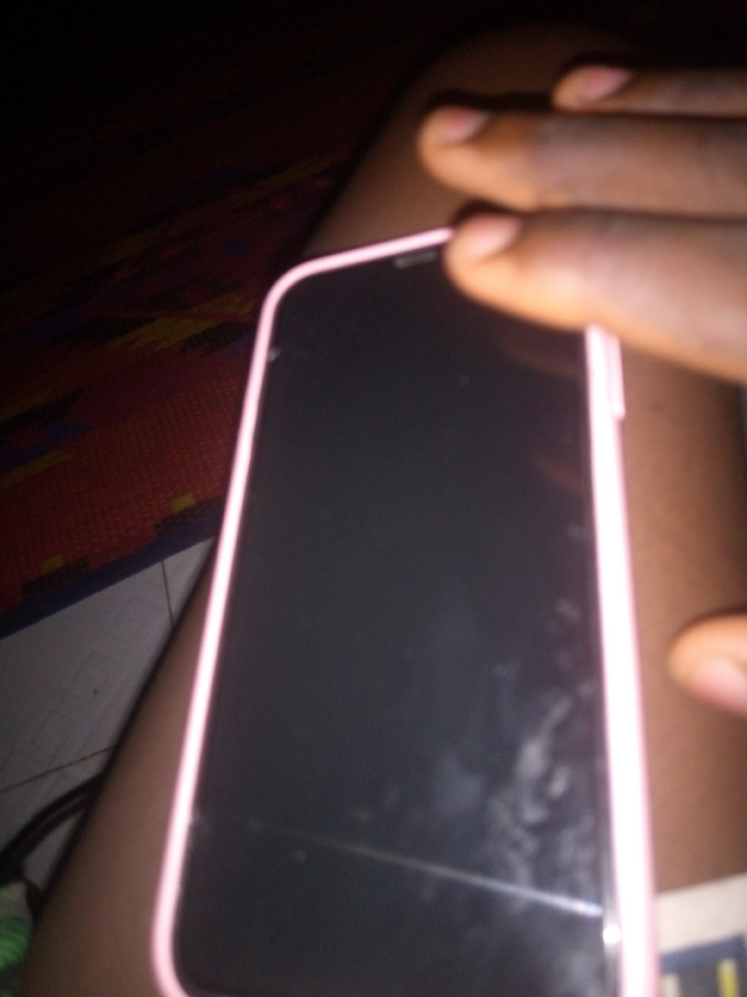 In an indoor setting with dim lighting, a person's hand, with visible fingernails and skin tone, is positioned over a smartphone. The smartphone has a pink casing and a dark screen. The background shows a textured surface, possibly a mat or carpet, with a red and yellow pattern. The overall image is out of focus, particularly the background and the hand. The location is Unknown, Unknown.