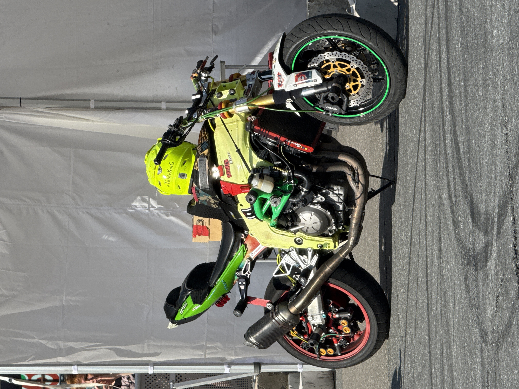 A neon yellow helmet rests on a custom-built motorcycle parked on asphalt. The motorcycle features a vibrant green tail section, a yellow frame, and a black exhaust pipe with a carbon fiber tip. The front wheel has a green rim sticker, and the rear wheel has a red rim and gold accents. The background consists of a large white tent or canopy, with a portion of a white railing and some people visible to the left. The scene appears to be set up for an event or exhibition, possibly a motorcycle show or competition. The asphalt surface shows signs of tire marks, suggesting recent activity. The location is not explicitly identified in the image, therefore it cannot be determined to be in Saint Petersburg, Russia.