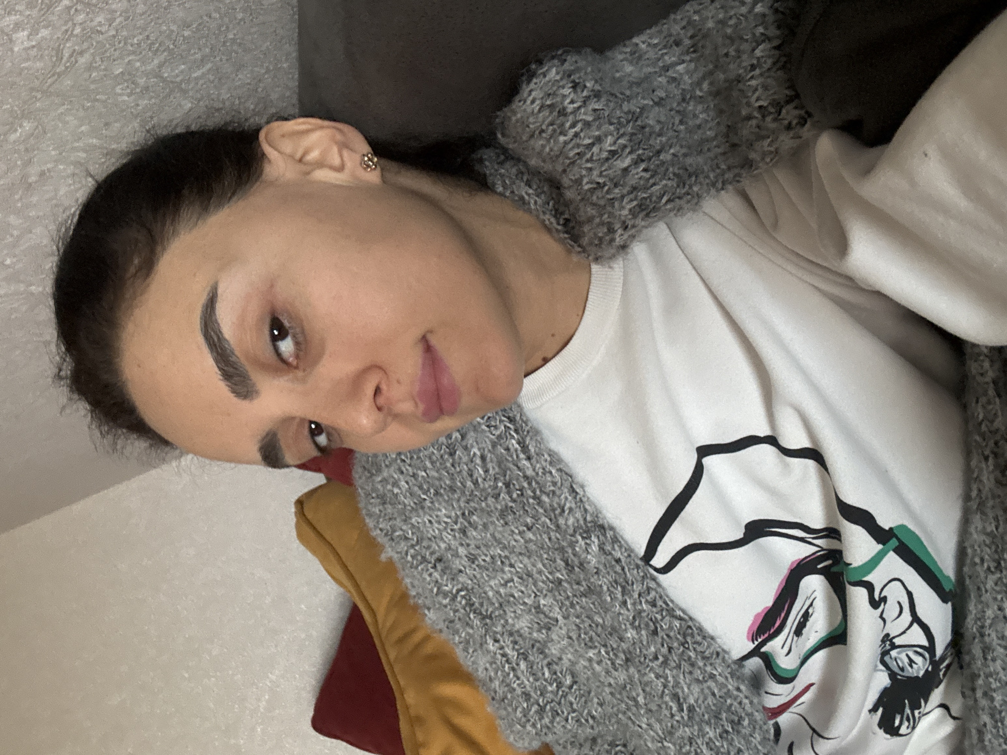 A woman is reclining indoors. She has dark hair pulled back, fair skin, and prominent eyebrows. She is wearing a white t-shirt with a graphic design on the front, and a gray knit scarf. She has an earring in her left ear. She is positioned on or near colorful pillows. The background appears to be a couch or wall. The image does not provide enough information to determine the location in Tbilisi, Georgia.