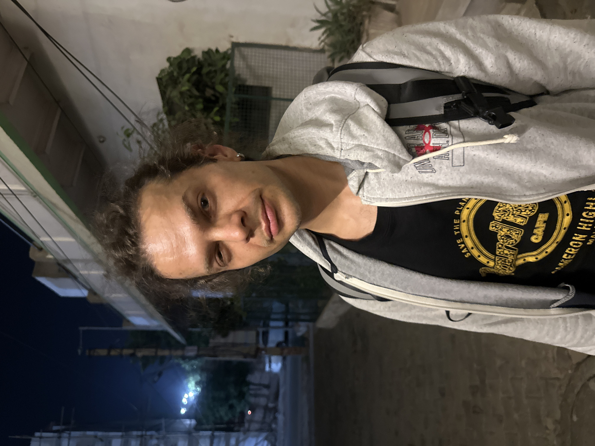 A man with curly hair, wearing a grey hoodie over a black T-shirt, stands outside at night. He is wearing a backpack and faces the camera. The background includes buildings, plants, and streetlights, suggesting an urban environment. The location is Mathura, India.