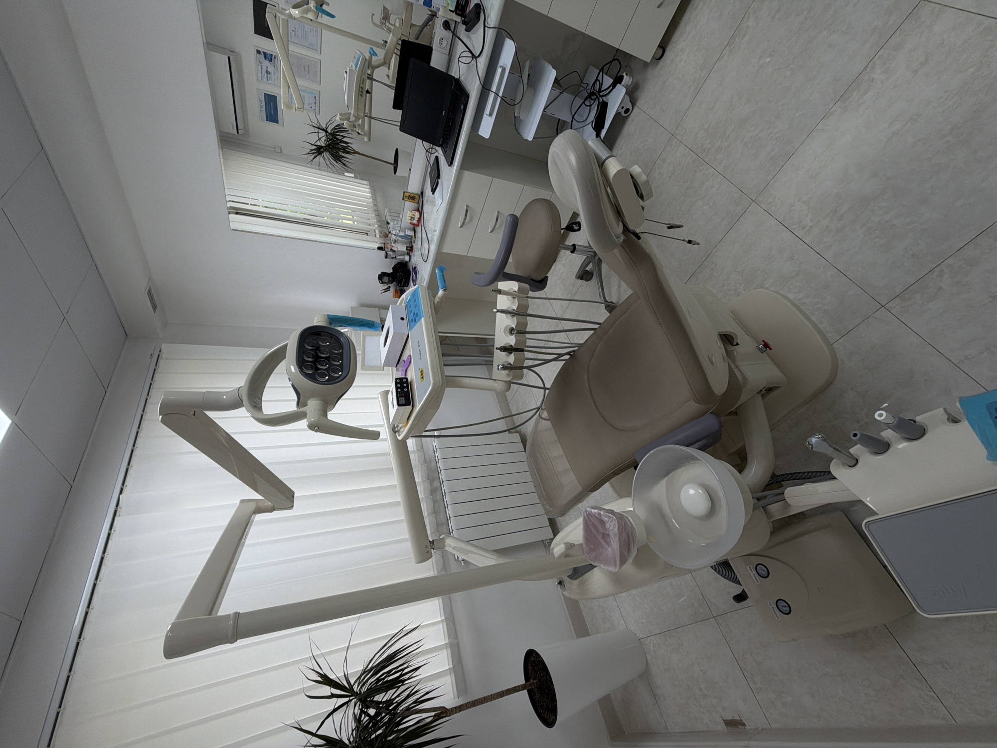 The image depicts a dental examination room in Tbilisi, Georgia. The room is brightly lit, with a dental chair as the central focus. The chair is beige and appears modern. To the left of the chair, a tall dental lamp is positioned, equipped with multiple LED bulbs. Next to the lamp, a plant in a white pot adds a touch of greenery to the clinical setting.
In the background, a desk is visible, holding a computer and other medical equipment. The walls are white, with diplomas or certificates hanging on the wall near the desk. The floor is covered in light-colored, square tiles. A dental assistant's chair is placed near the main chair. The room appears clean and well-maintained.