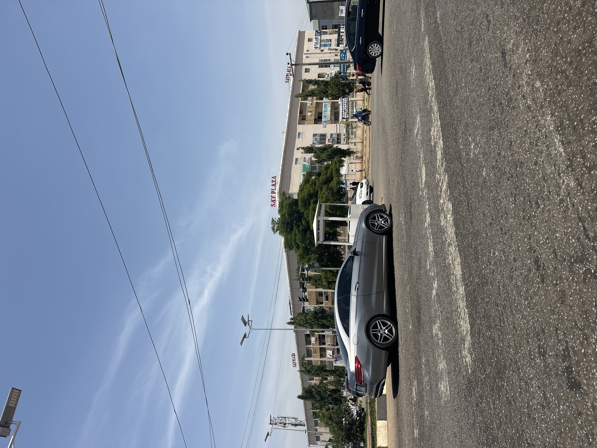 The image depicts a street scene in Abuja, Nigeria, specifically in front of the "Say Plaza" building. The plaza is a multi-story commercial building with various signs for businesses visible. In the foreground, a silver Mercedes-Benz coupe is parked on the road. Several other vehicles, including a black sedan and a motorcycle, are also present on the street, suggesting active traffic. Pedestrians can be seen walking near the plaza and crossing the street. A white traffic officer booth is also visible. The sky is clear with some wispy clouds, indicating a sunny day. The overall environment appears to be a busy urban area with commercial activity.